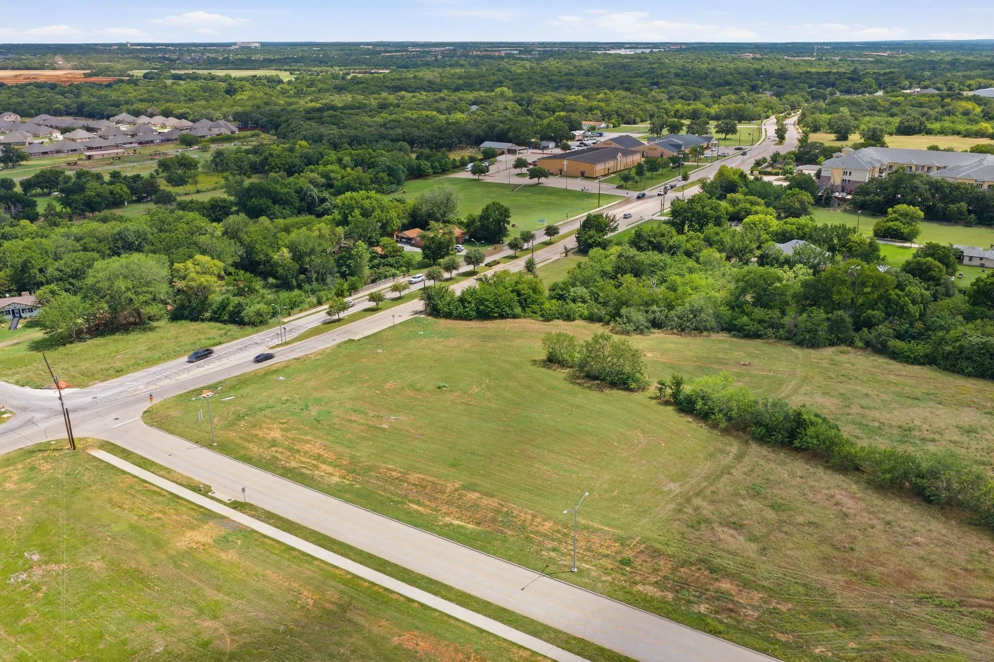 Expansive land parcel featuring a paved road frontage, sidewalk infrastructure, and surrounding mature trees