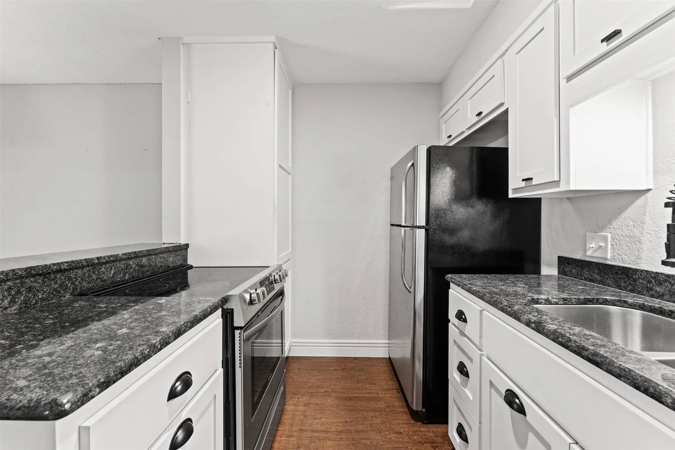 Galley kitchen featuring dark granite countertops, white cabinetry with dark hardware, stainless steel appliances, and wood-finish flooring