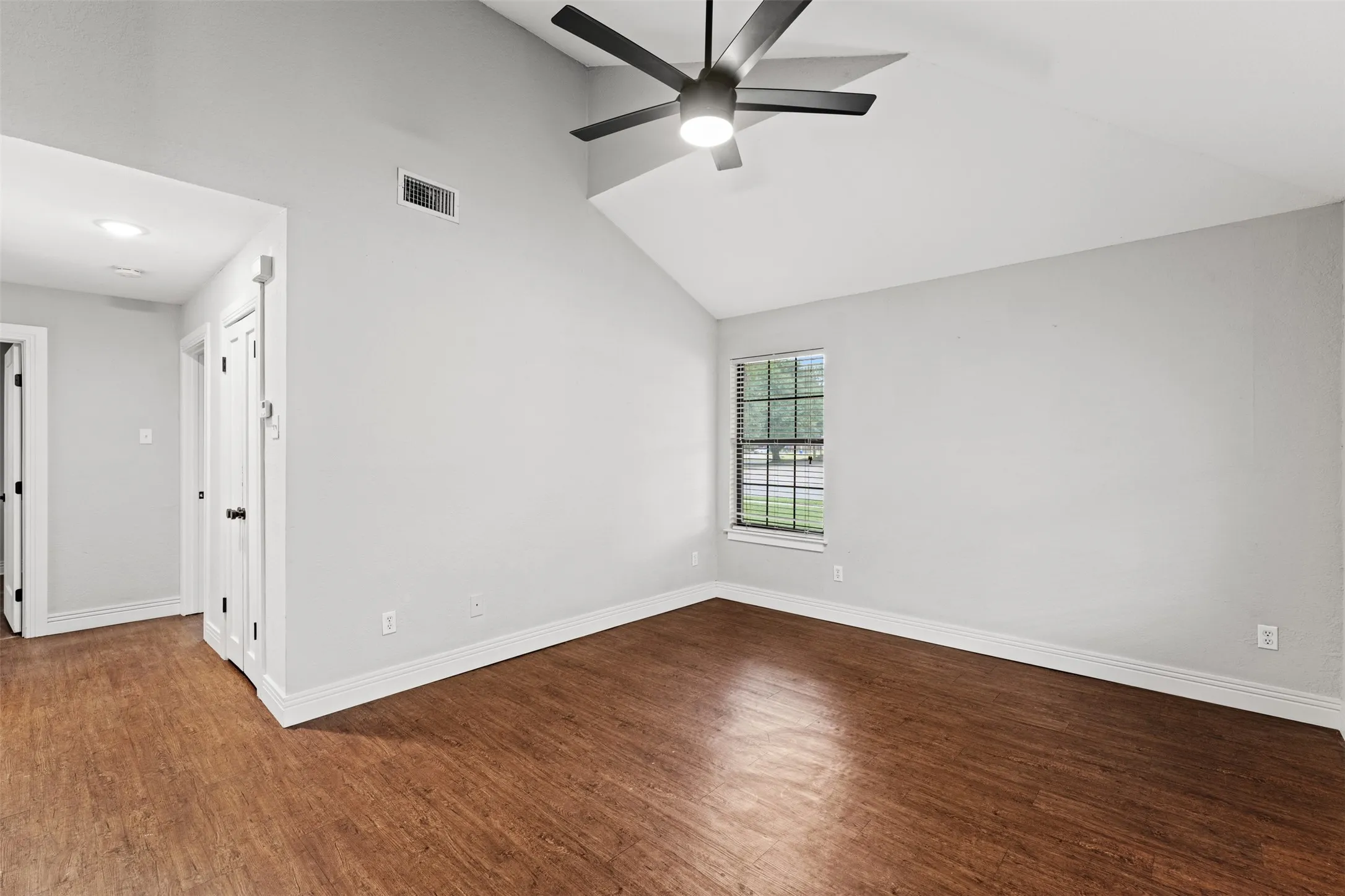Vaulted ceiling room featuring wood-finish flooring, a contemporary ceiling fan with integrated lighting, window with blinds, and white trim
