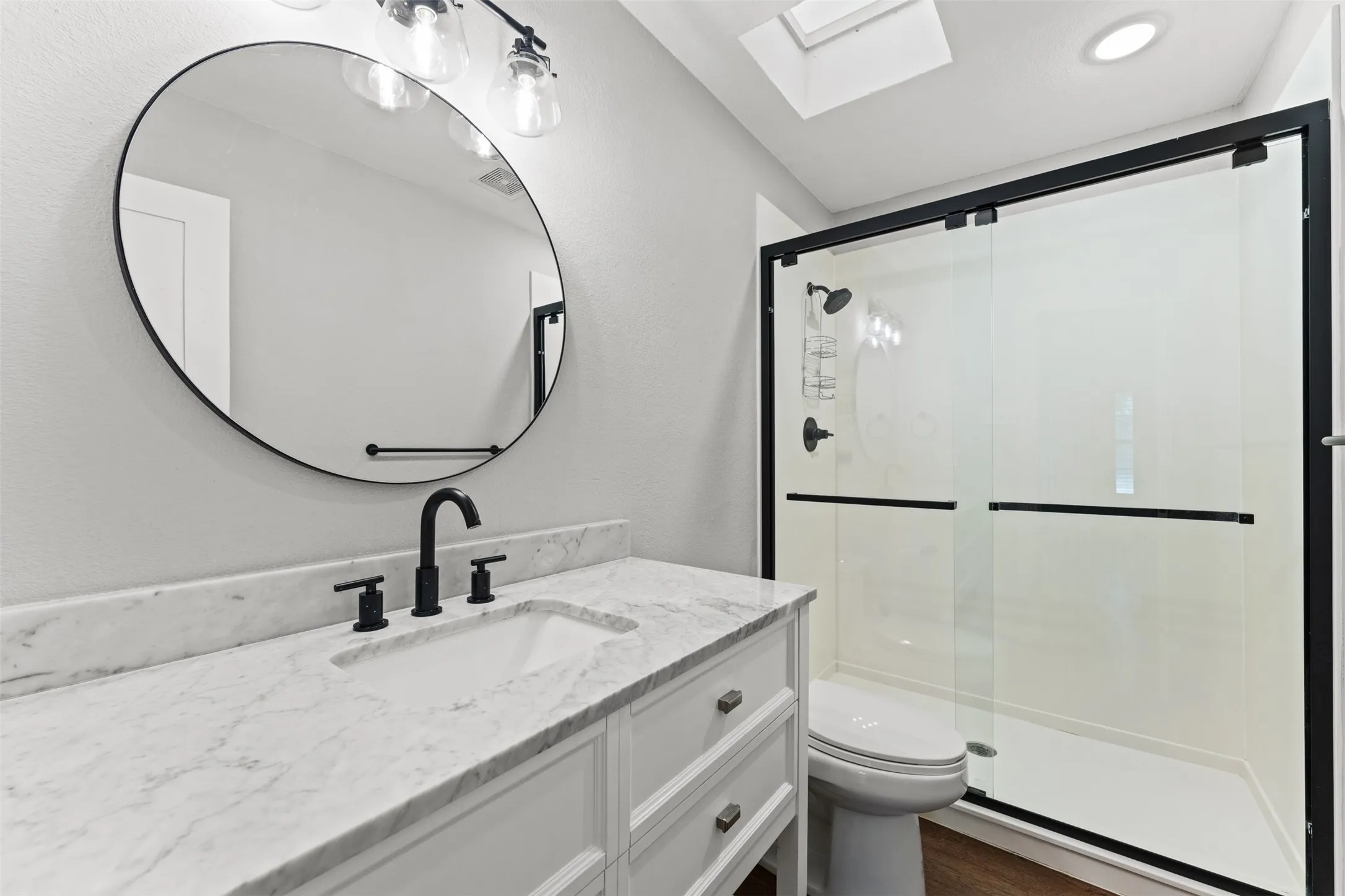 Bathroom featuring a white vanity with a stone countertop, a round frameless mirror, matte black fixtures, and a glass-enclosed shower with a black frame