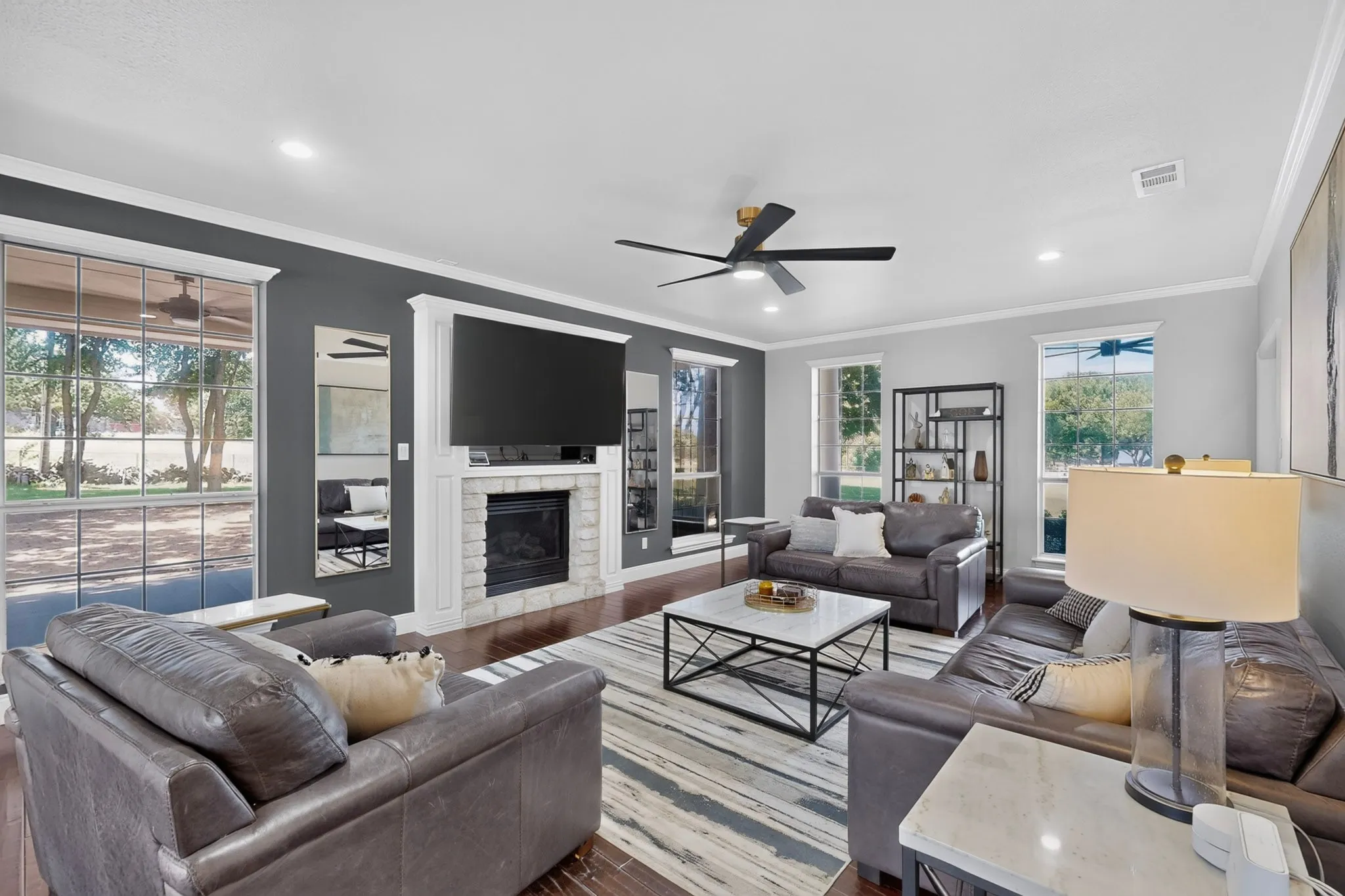 Living room featuring a stone-clad fireplace with a white mantel, rich wood-finish flooring, recessed lighting, a ceiling fan, and multiple windows