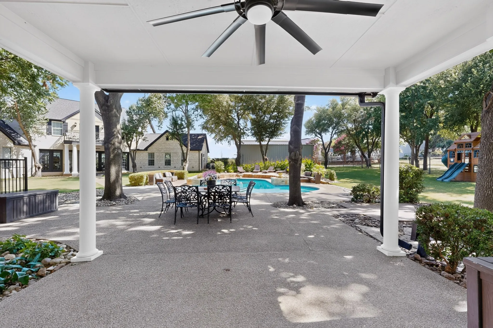 Covered patio with an exposed aggregate concrete surface, featuring a ceiling fan, white support columns, and lush landscaping