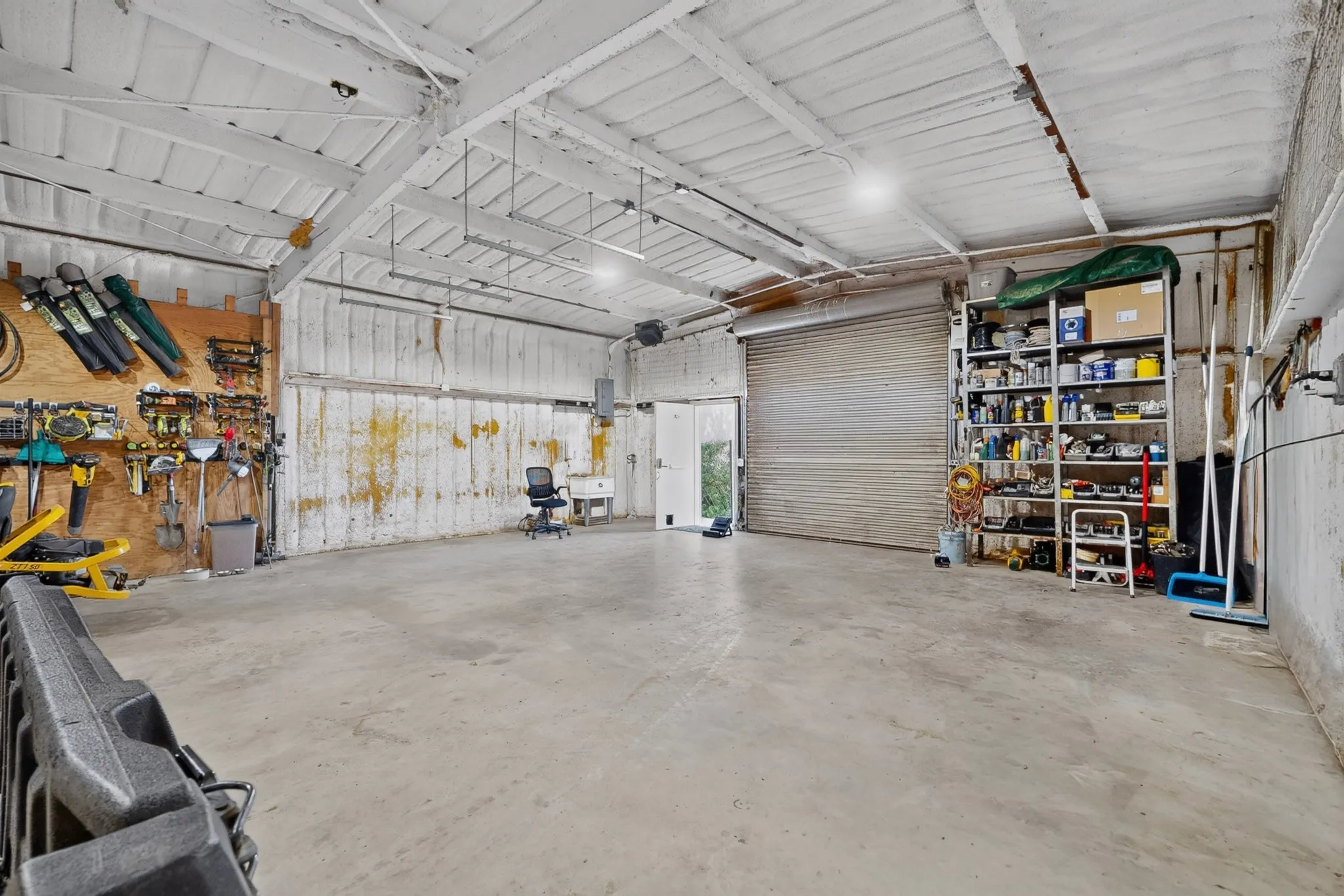 Spacious interior featuring a large roll-up door, concrete flooring, exposed beam ceiling, built-in shelving, and a utility sink