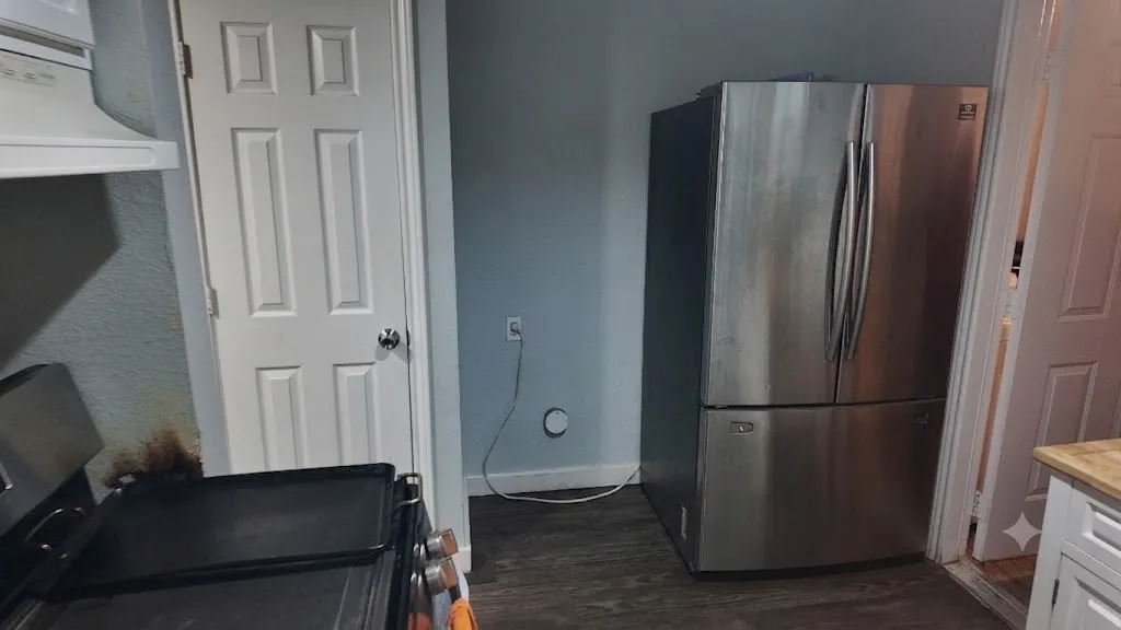 Stainless steel French door refrigerator set against a light blue wall, featuring dark wood-finish flooring and a white paneled door