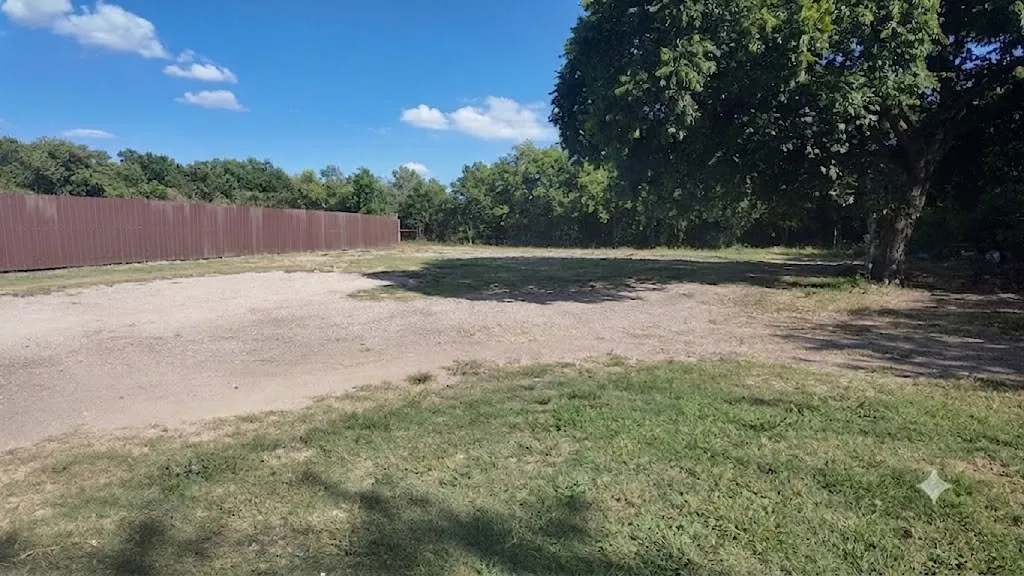 Expansive lot featuring a substantial brown privacy fence, mature trees, a gravel area, and natural ground cover