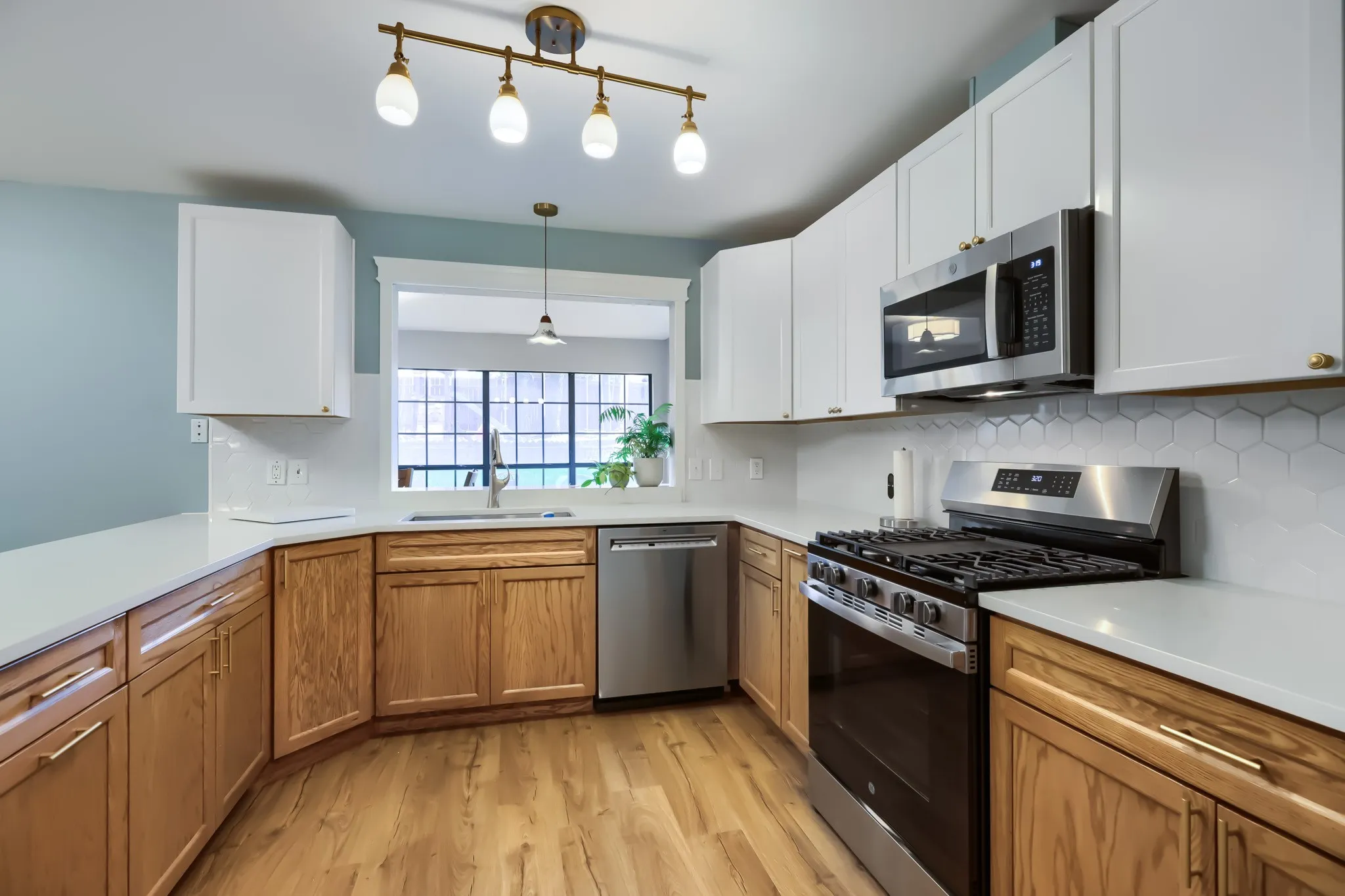 Modern kitchen featuring wood-finish flooring, white upper cabinetry, natural wood lower cabinetry, light-toned countertops, and stainless steel appliances including a range and microwave
