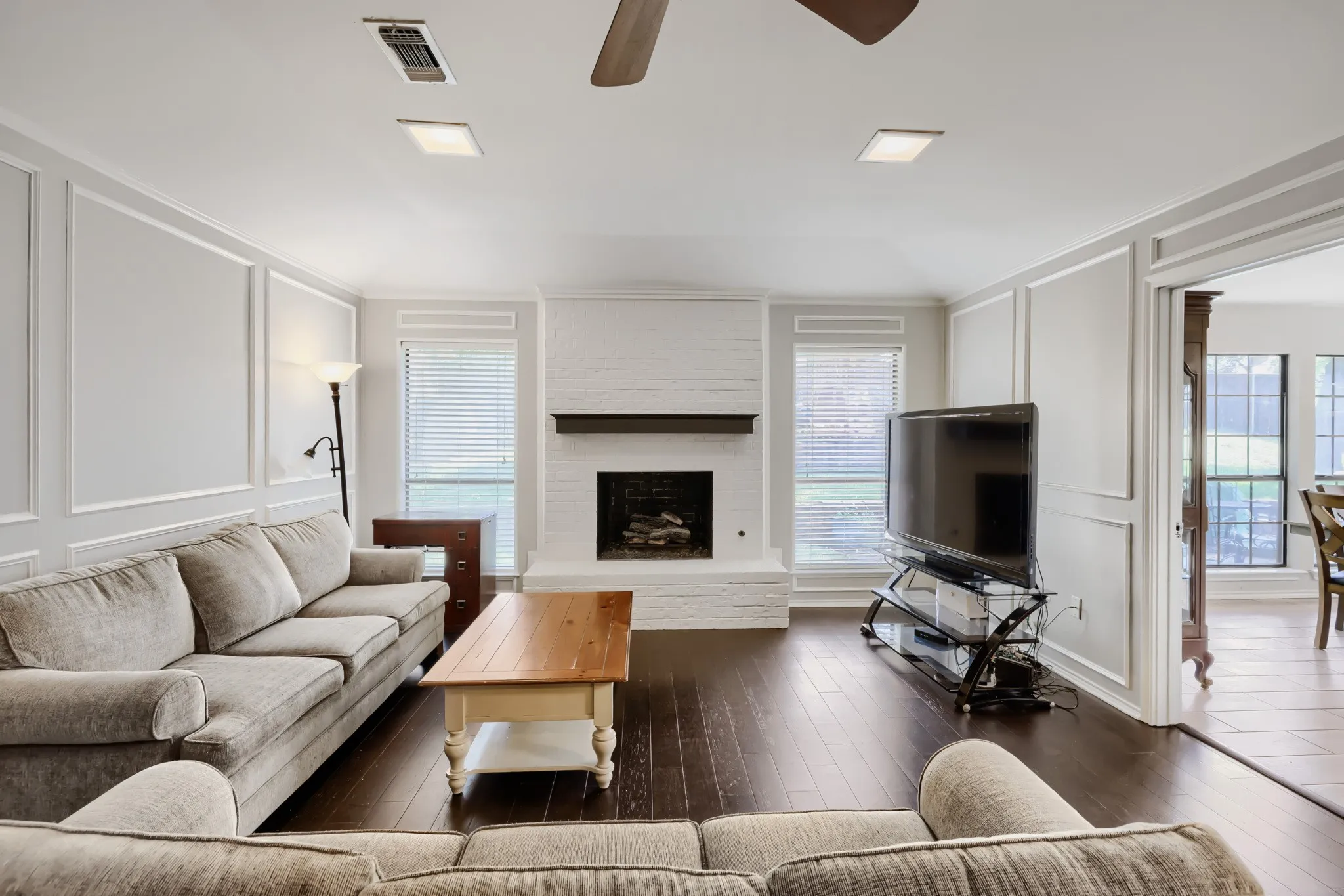 Spacious living area featuring dark wood-finish flooring, white brick fireplace with a wood mantle, and decorative wall molding