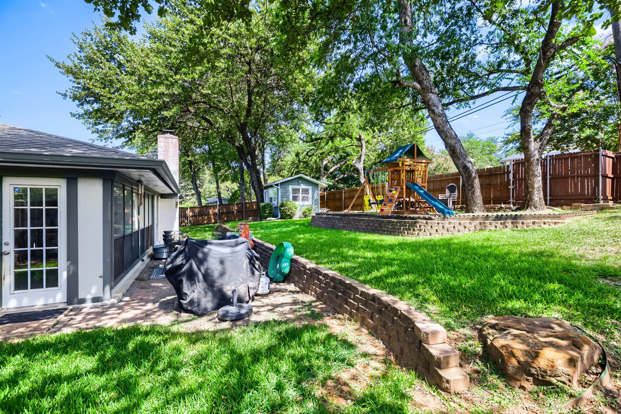 Spacious backyard featuring a brick patio, mature trees, a wood privacy fence, and a tiered lawn with a brick retaining wall