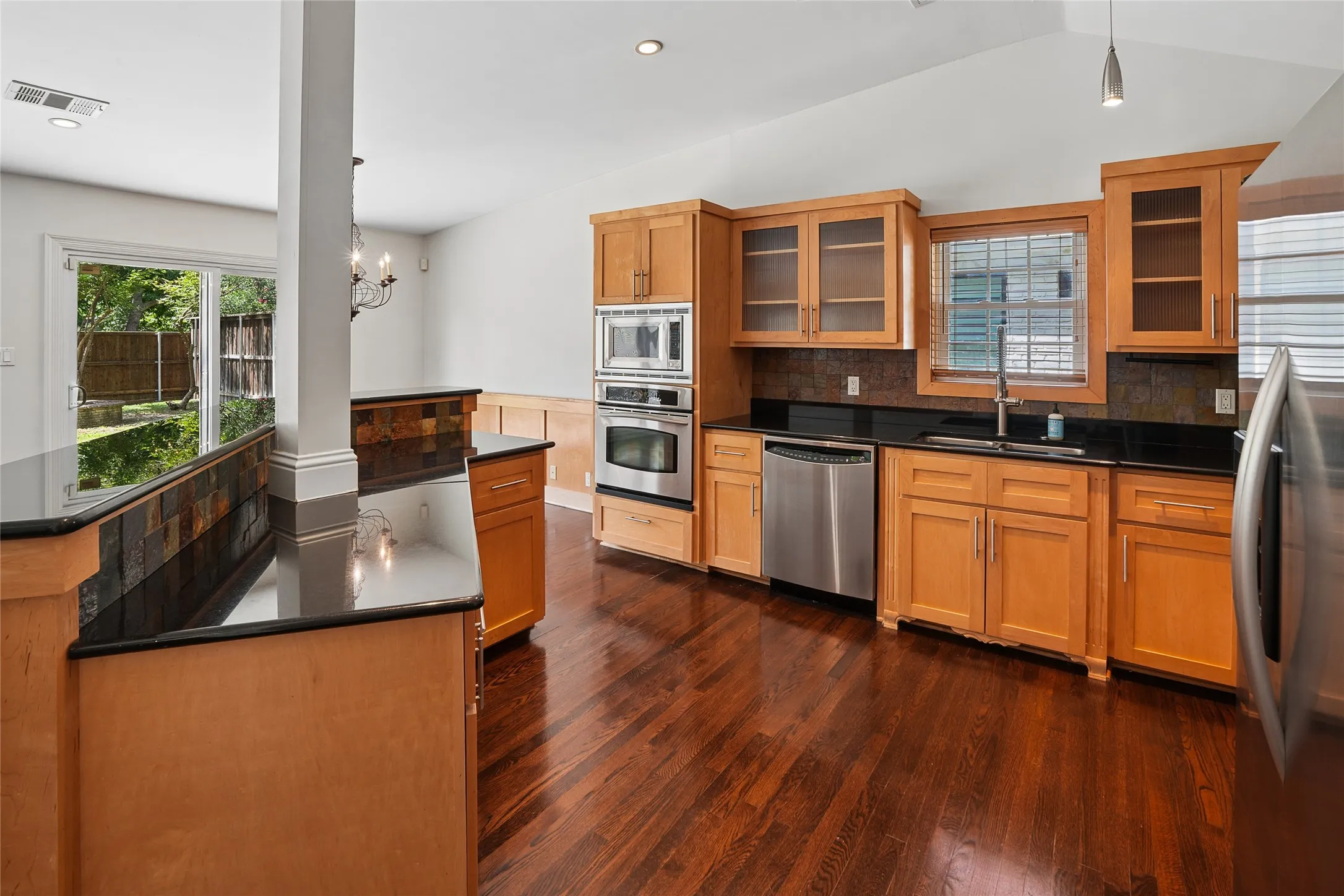 Kitchen featuring wood-finish flooring, light wood cabinetry, dark countertops, built-in stainless steel appliances, and a tile backsplash