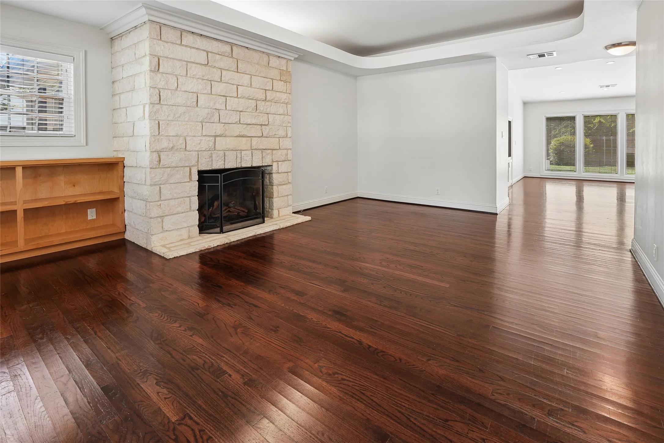 Living area featuring rich wood-finish flooring, a stone fireplace with hearth, built-in shelving, white trim, and tray ceiling details
