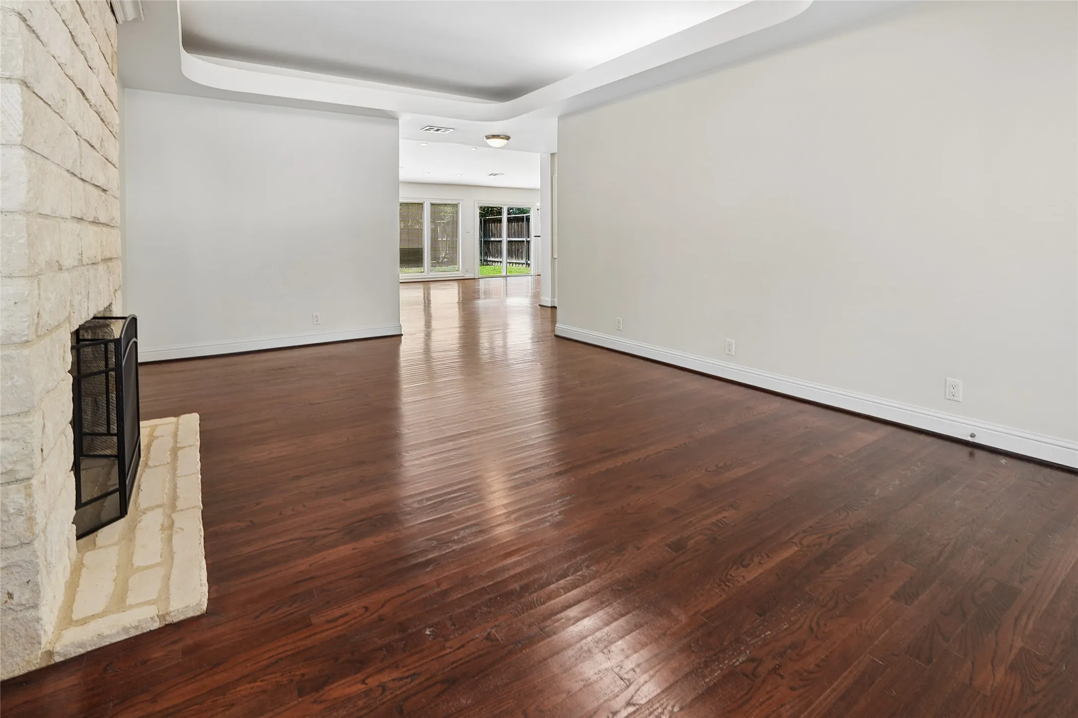 Spacious room featuring rich wood-finish flooring, a white brick fireplace with a hearth, white baseboards, and a tray ceiling