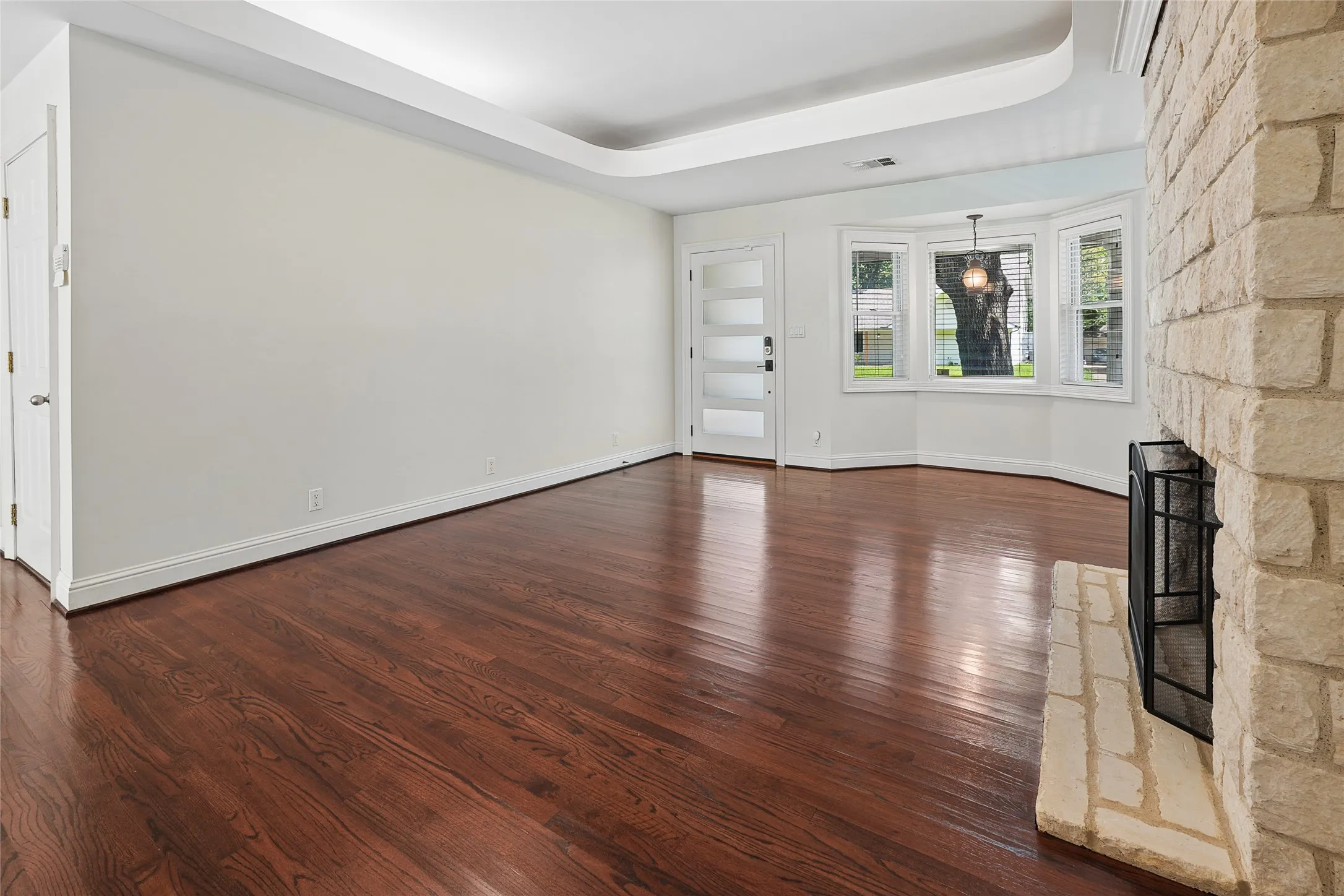 Spacious interior featuring rich wood-finish flooring, a stone-clad fireplace with a hearth, a tray ceiling, and a bay window with natural light