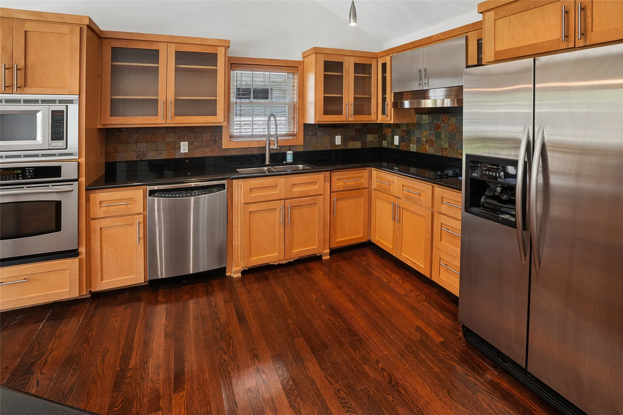 Kitchen featuring wood-finish flooring, light wood cabinetry, black countertops, and a tile backsplash