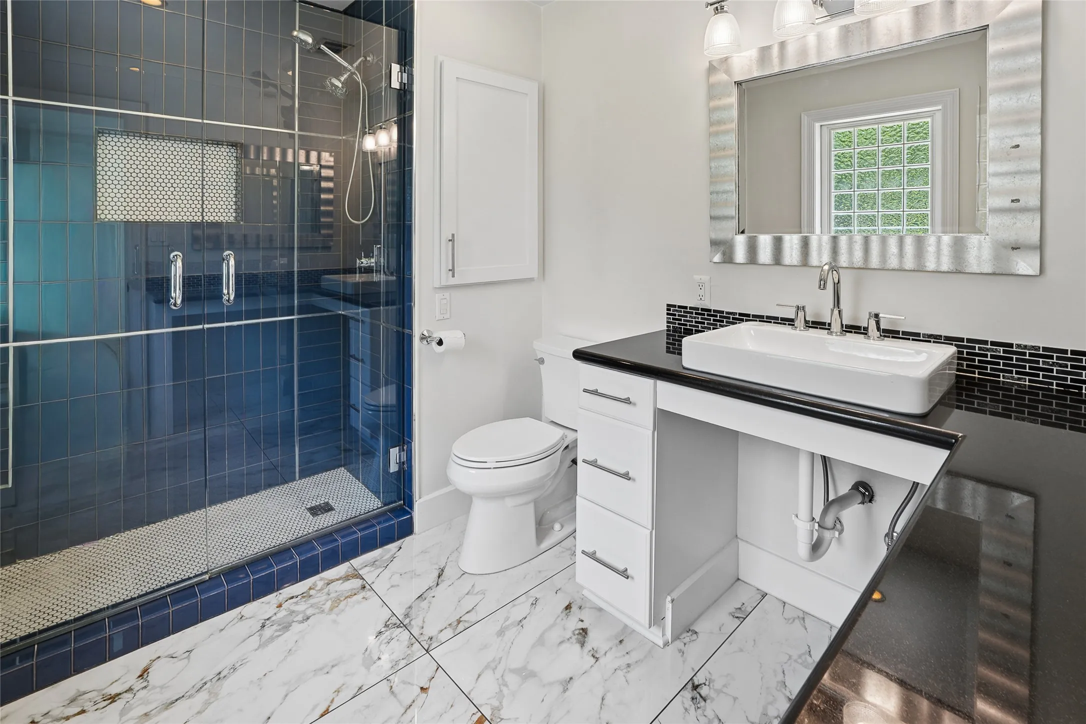 Contemporary bathroom featuring a glass-enclosed shower with blue subway tile, a white vessel sink on a dark countertop, an elegant vanity mirror, and white-and-gray patterned floor tiling