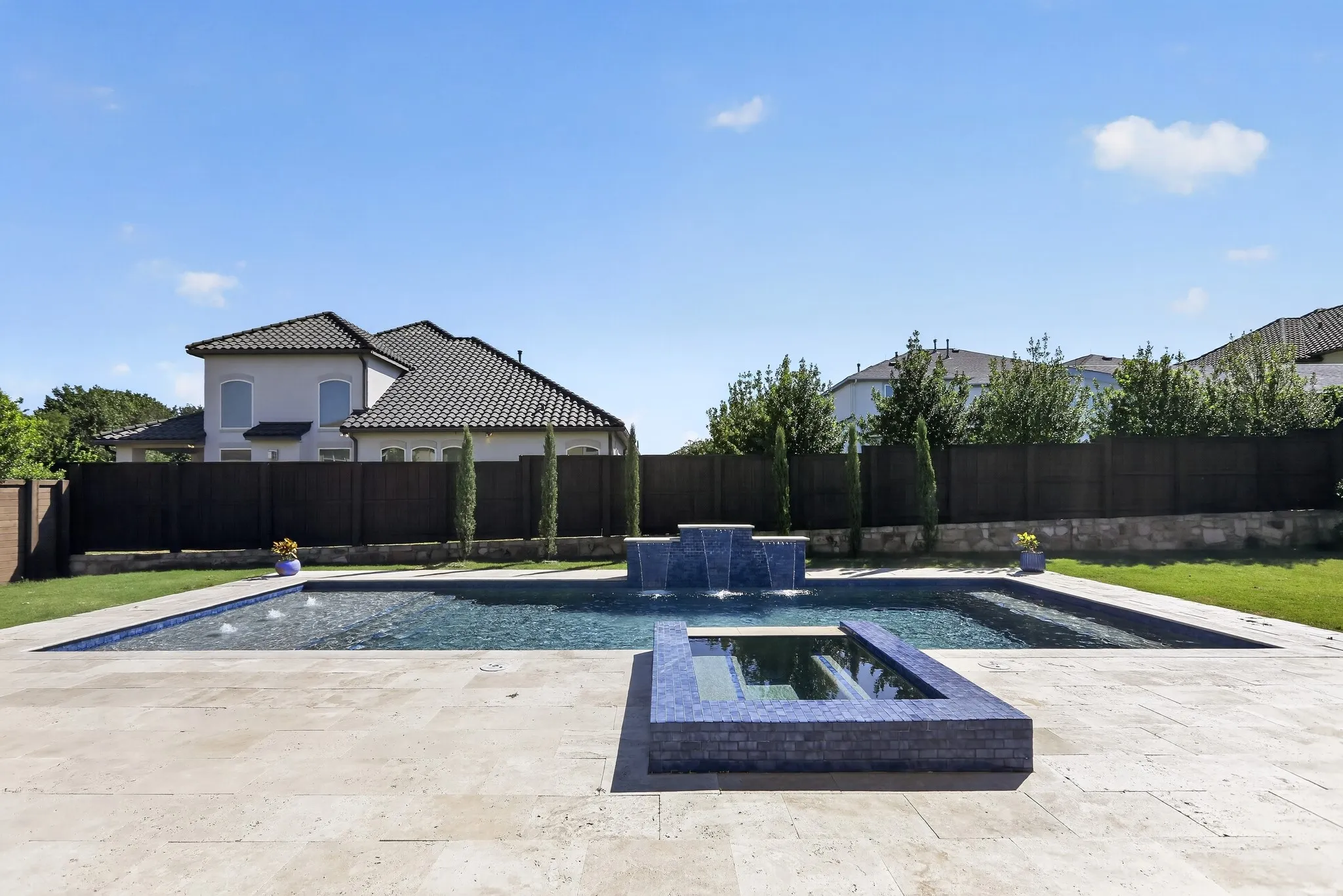 Outdoor living space featuring a rectangular swimming pool with integrated spa and water features, surrounded by a light-toned stone patio