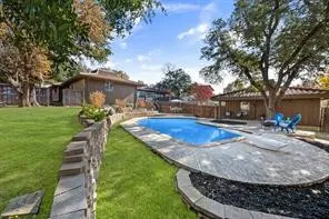 Backyard oasis featuring an in-ground pool with a stone surround, an adjacent covered patio structure, a stacked stone retaining wall, and expansive lawn areas