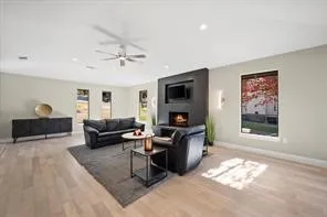 Spacious living area featuring light wood-finish flooring, a modern black fireplace surround with an integrated media niche, recessed lighting, a ceiling fan, and multiple windows