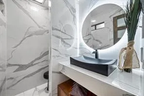 Modern bathroom featuring white walls and flooring with gray veining, a floating vanity with a vessel sink, a matte black faucet, and a back-lit circular mirror
