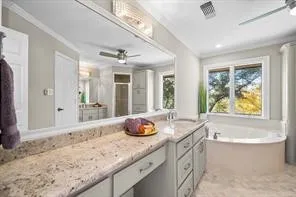 Spacious bathroom featuring a large soaking tub, double window, granite-style countertop, extensive cabinetry, and a ceiling fan