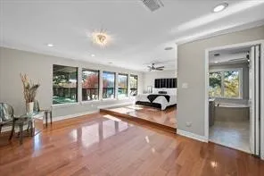Expansive room featuring rich wood-finish flooring, a raised platform with additional wood-finish flooring, a wall of multiple windows, recessed ceiling lighting, and an adjacent room with a built-in tub and window