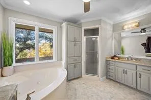 Soaking tub with surrounding window, tile flooring, glass-enclosed shower, light-colored cabinetry, and dual vanity with integrated sinks