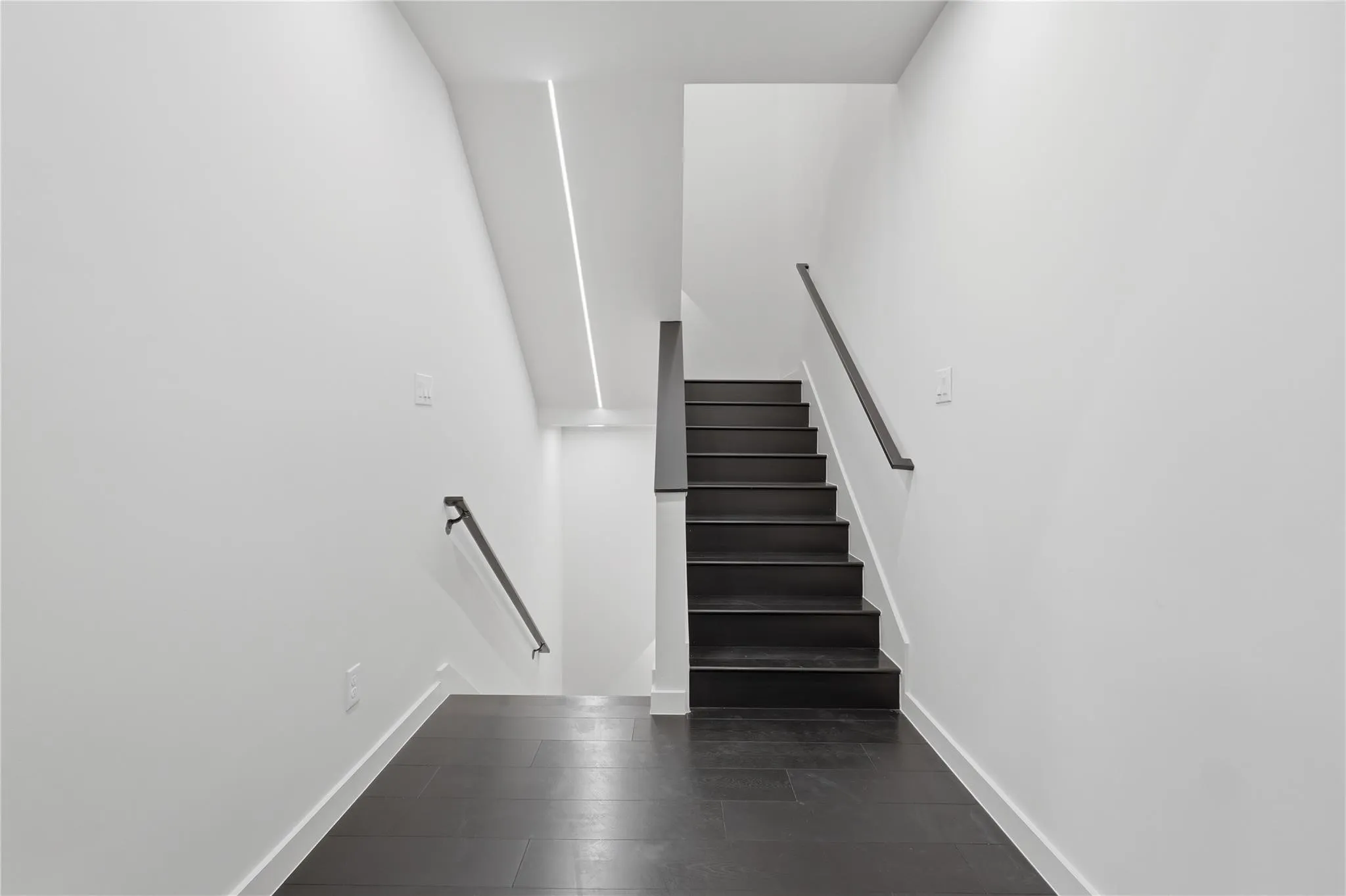 Stairwell featuring dark wood-finish flooring and stairs, white walls, black handrails, and recessed linear ceiling lighting