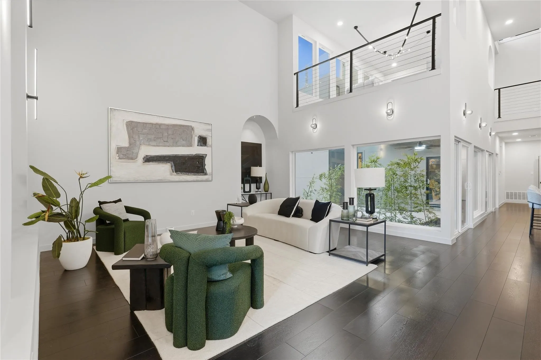 Double-height living space featuring dark wood-finish flooring, a contemporary staircase with cable railing, large windows with exterior foliage, and recessed lighting