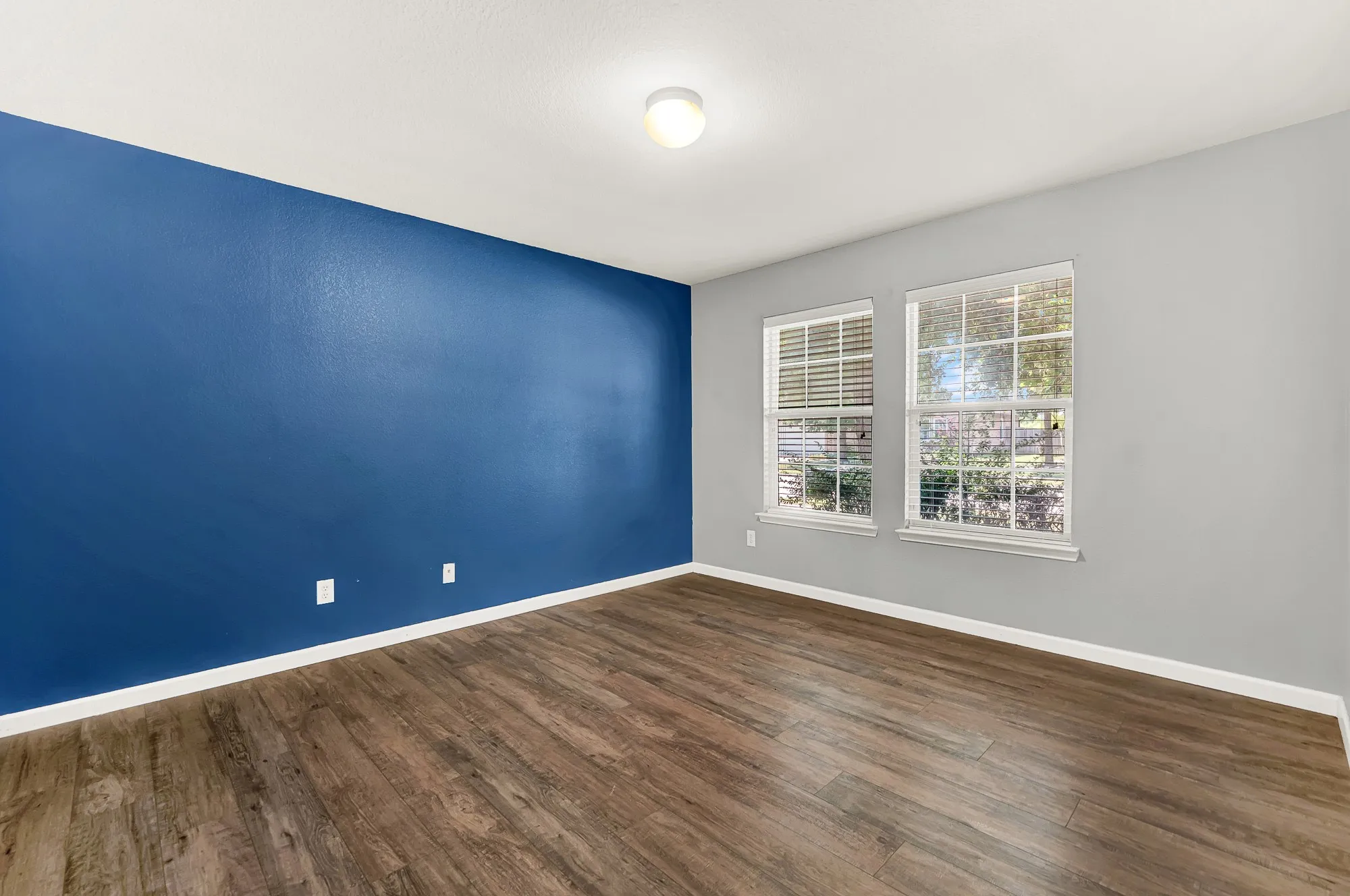 Spacious room featuring wood-finish flooring, a vibrant accent wall, two large windows with blinds, and recessed lighting