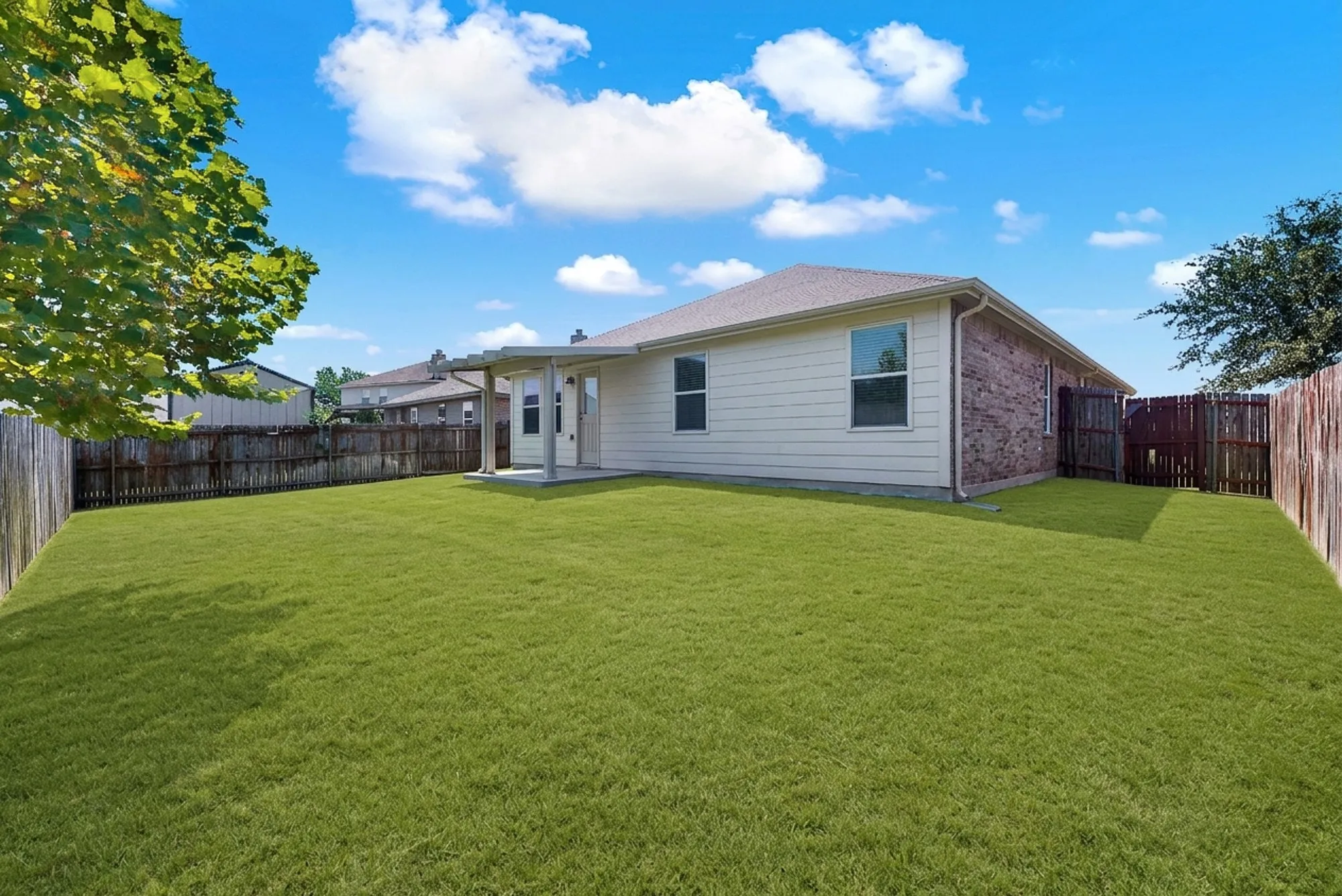 Expansive grass yard bordered by a wood fence, featuring a covered concrete patio with support columns, and a house exterior with light siding and brick accents