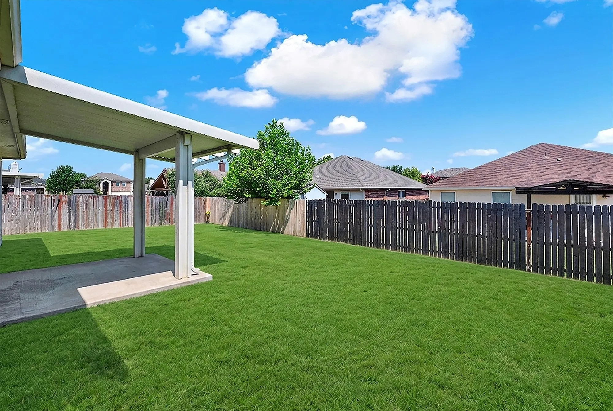 Expansive backyard featuring a covered patio with concrete flooring, a lush green lawn, and a privacy fence