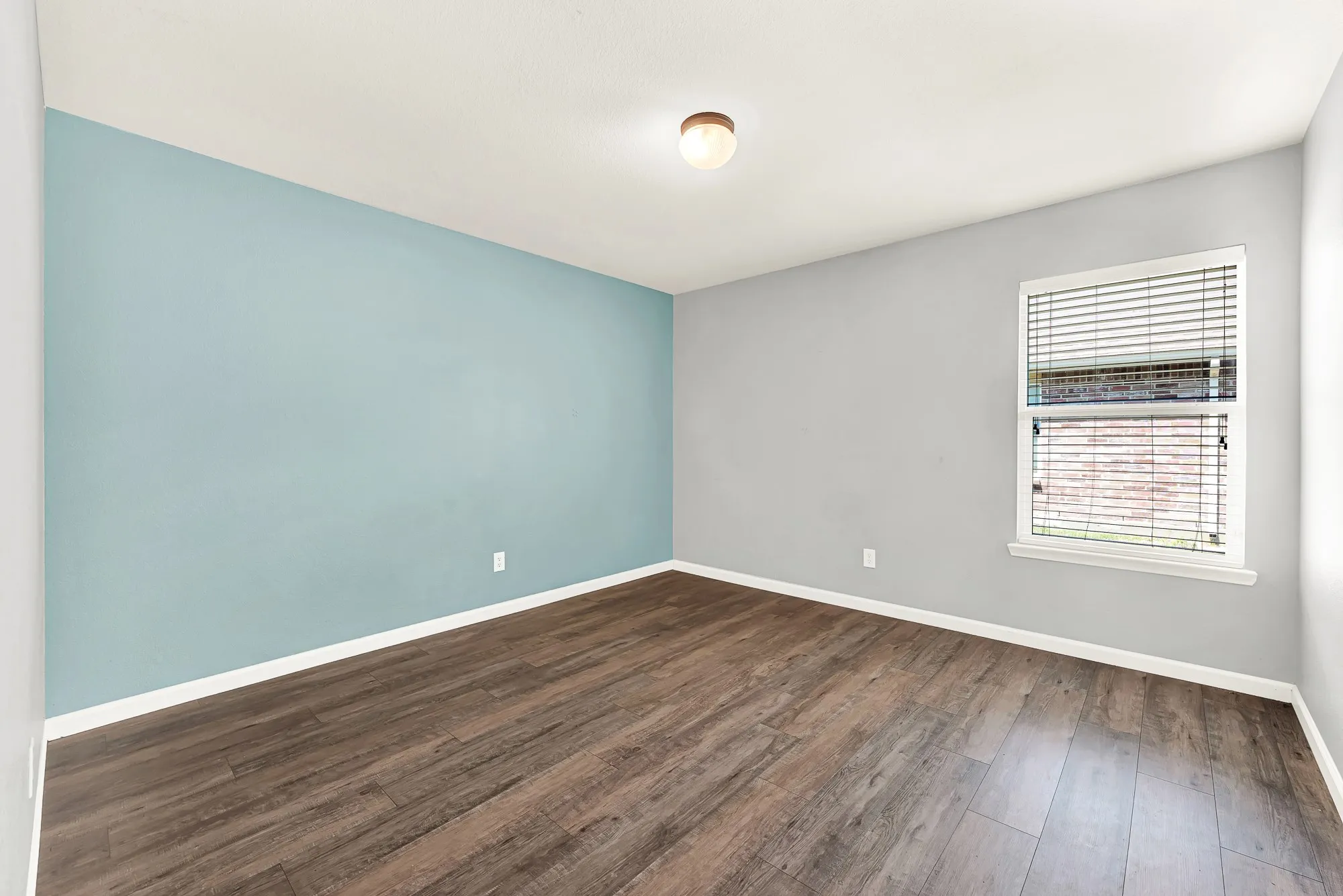 Room featuring wood-finish flooring, a light blue accent wall, and a single window with horizontal blinds