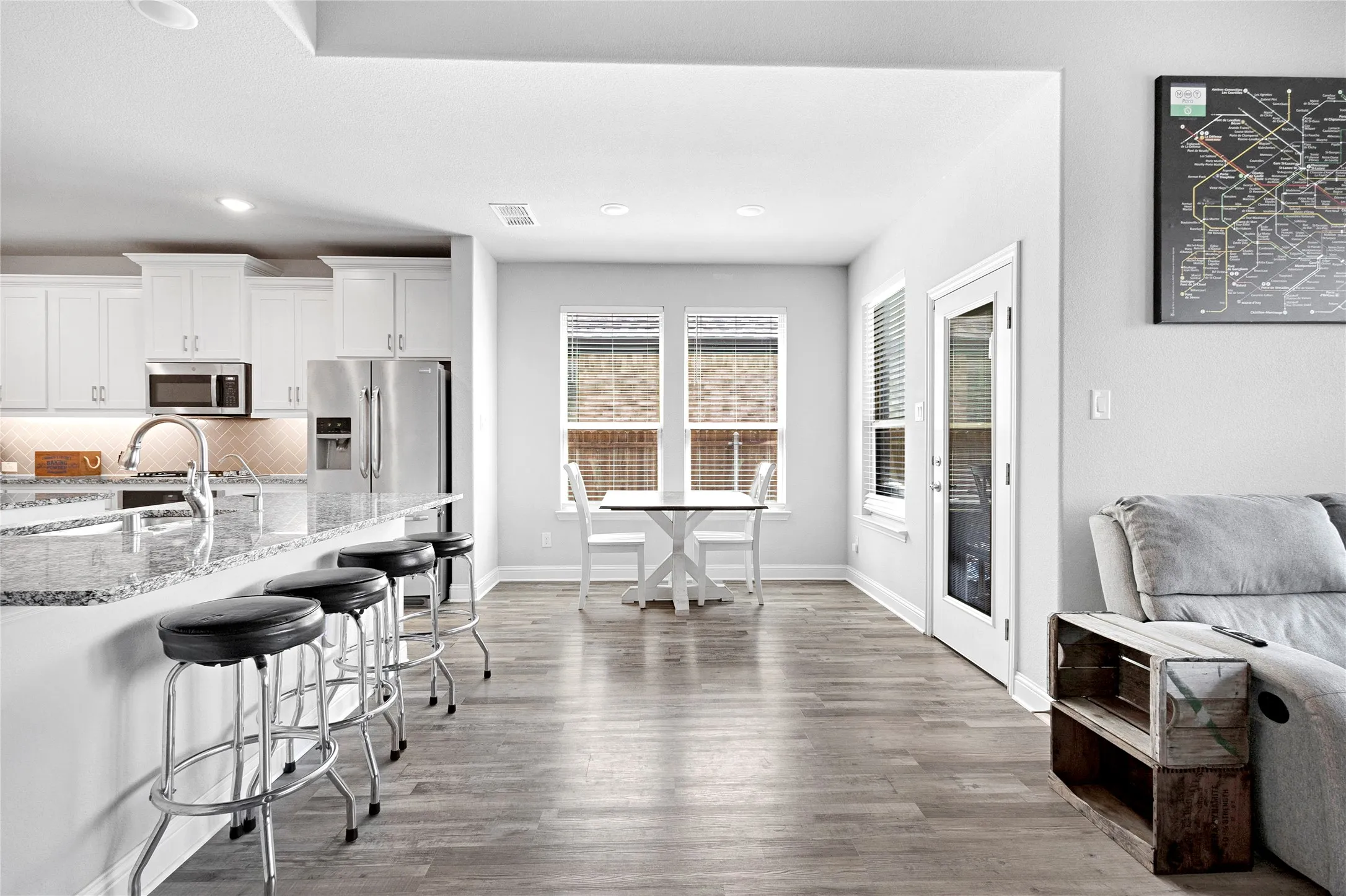 Open-concept living space featuring wood-finish flooring, a kitchen island with seating, white cabinetry, stainless steel appliances, and ample natural light from multiple windows