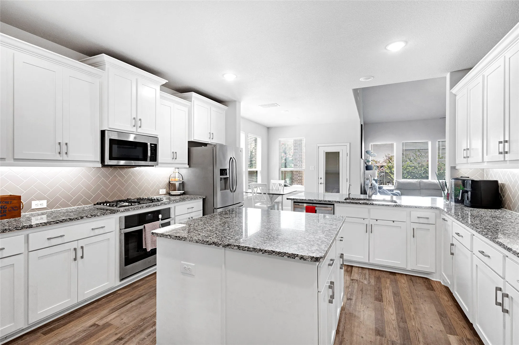 Modern kitchen featuring white cabinetry, granite countertops, stainless steel appliances, wood-finish flooring, and a herringbone tile backsplash