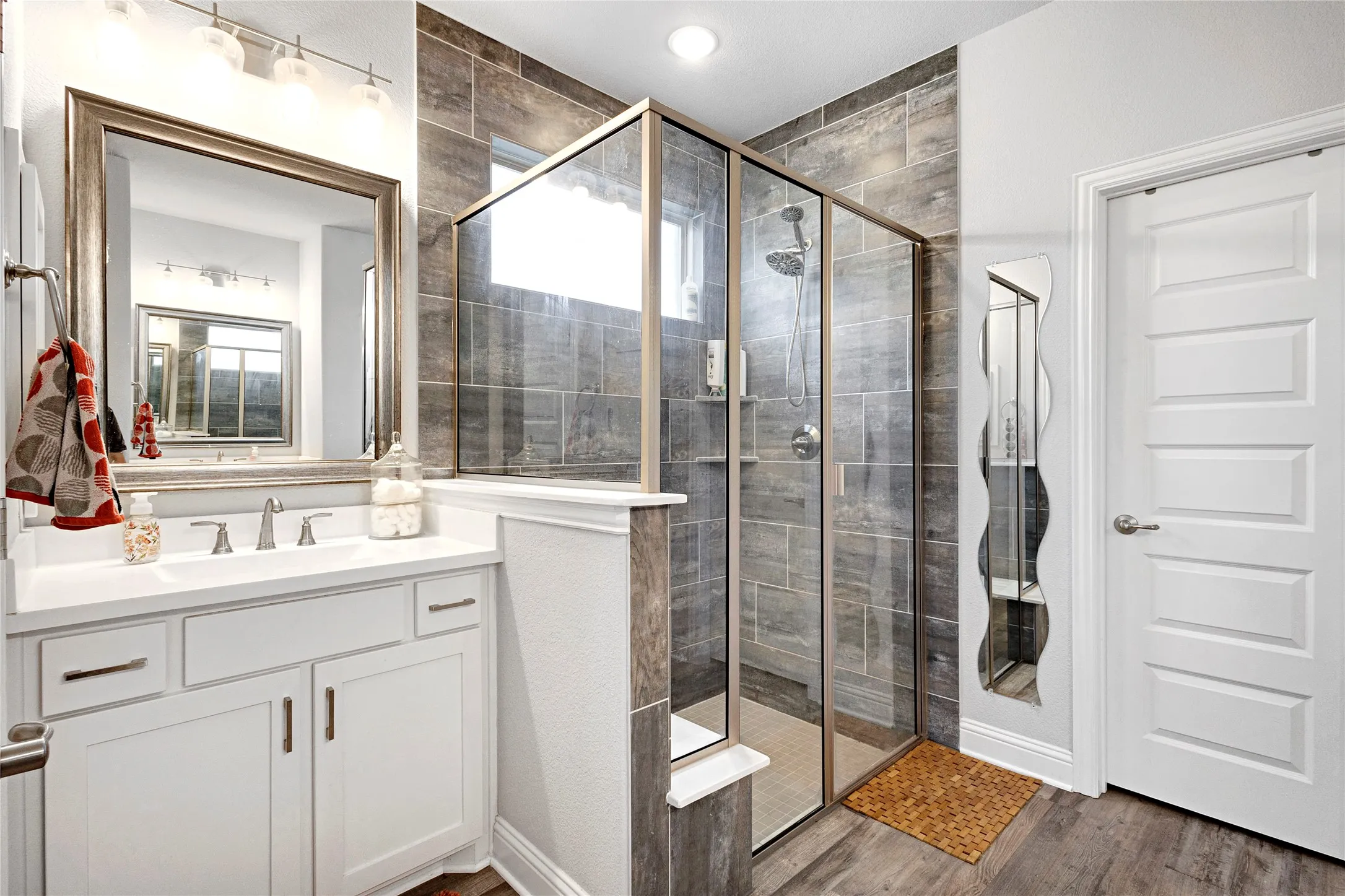 Contemporary bathroom featuring a glass-enclosed shower with tile surround, a white vanity with integrated sink, a framed mirror, and wood-finish flooring