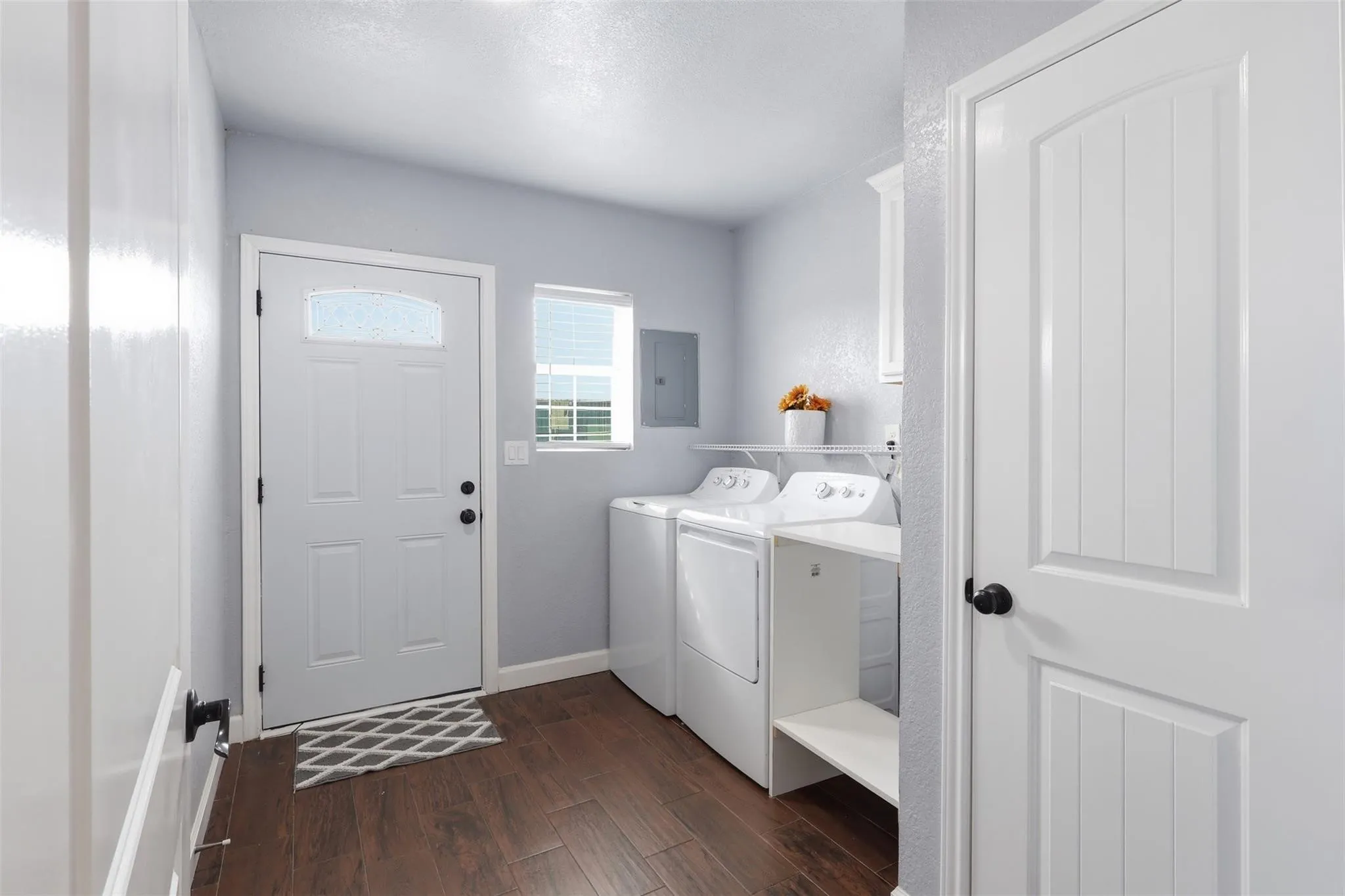 Laundry area featuring wood-finish flooring, light gray walls, a window with blinds, a side-entry door with an arched glass insert, and built-in white shelving