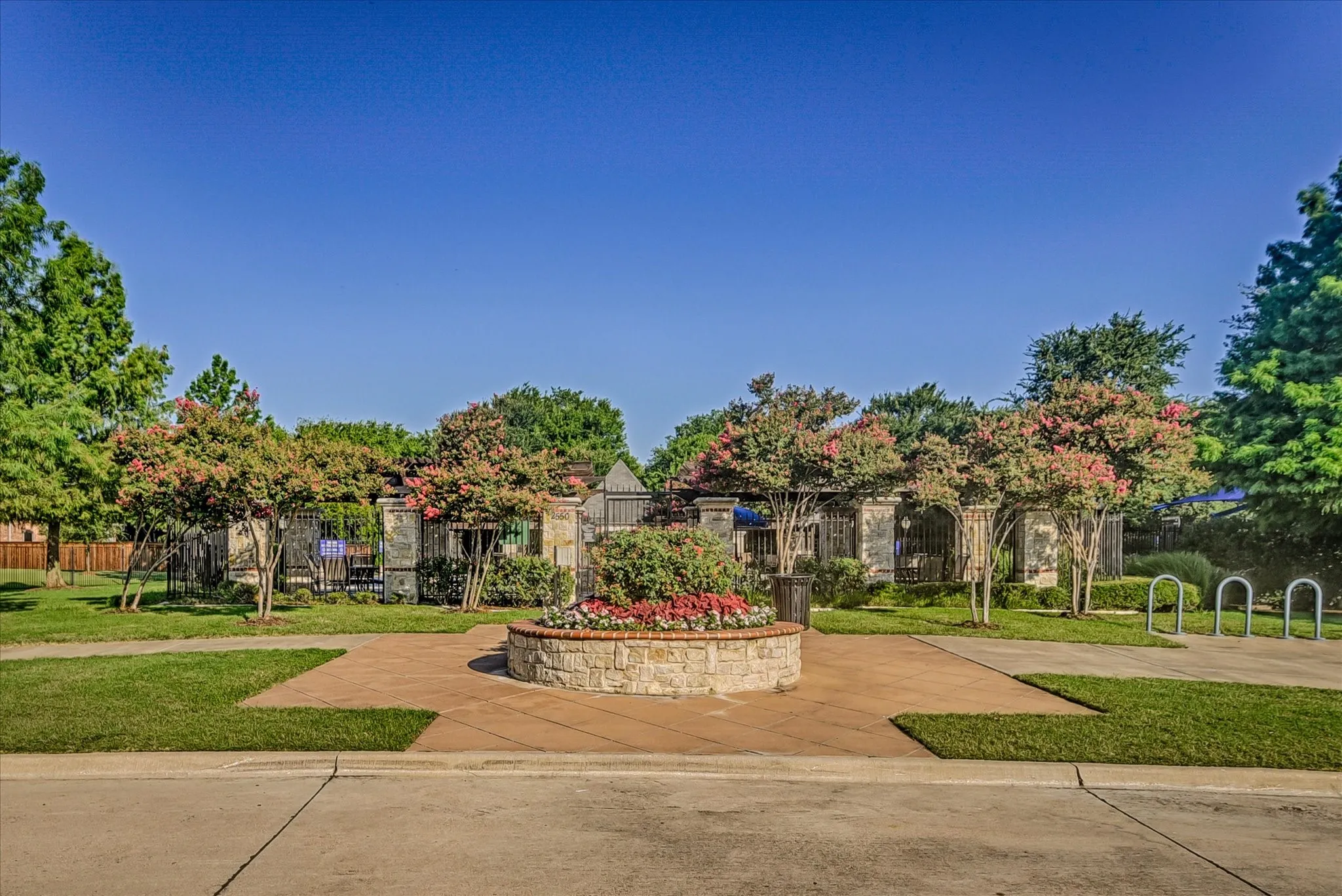 Community amenity area featuring a gated entrance, stone-clad columns, mature landscaping with flowering trees, a decorative stone planter, and bike racks
