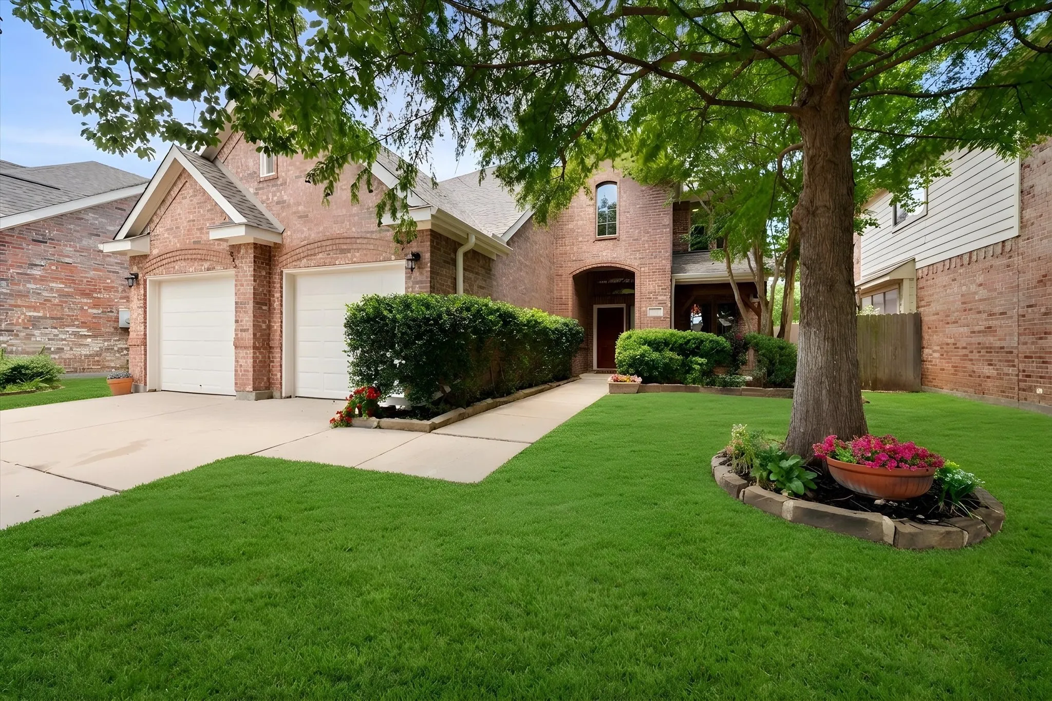Brick facade home featuring a two-car garage, mature front yard tree, established shrubbery, and a concrete pathway leading to the entry