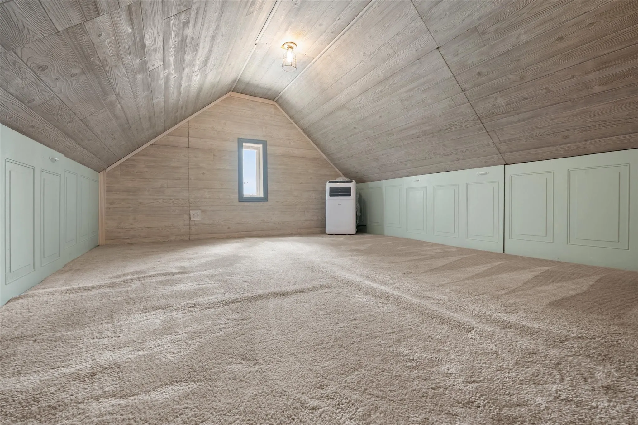 Spacious garage attic area featuring AC, sloped wood-finish paneling, light-toned carpeting, and a framed window