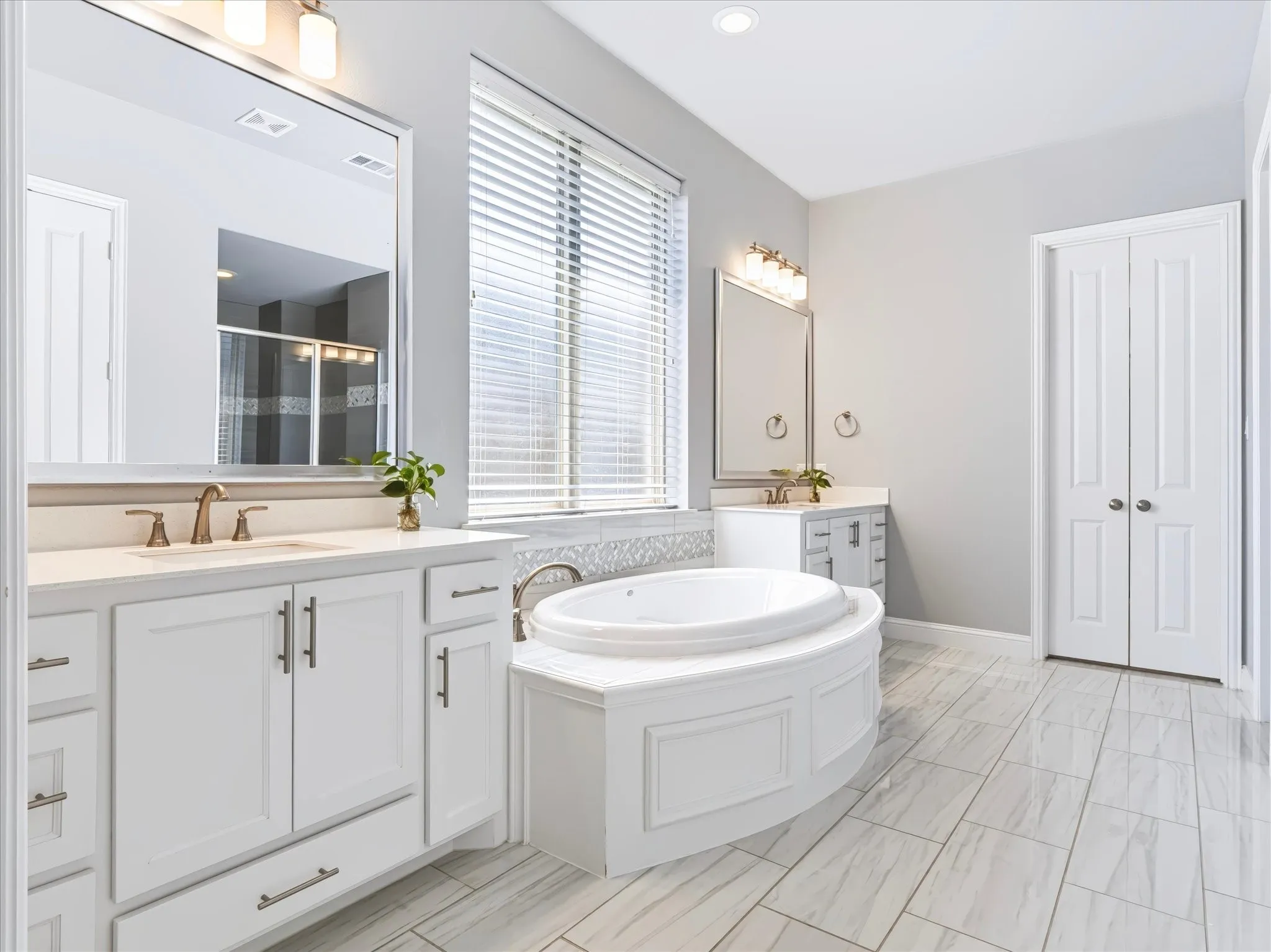 Double vanity with white cabinetry and integrated sinks, a large soaking tub with tiled surround, and white tile flooring