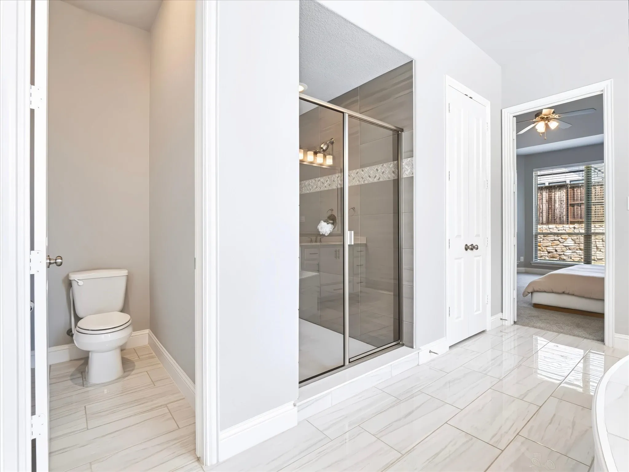 Spacious bathroom featuring large format tile flooring, a glass-enclosed shower with tile surround, a built-in toilet nook, and a dual vanity with a framed mirror