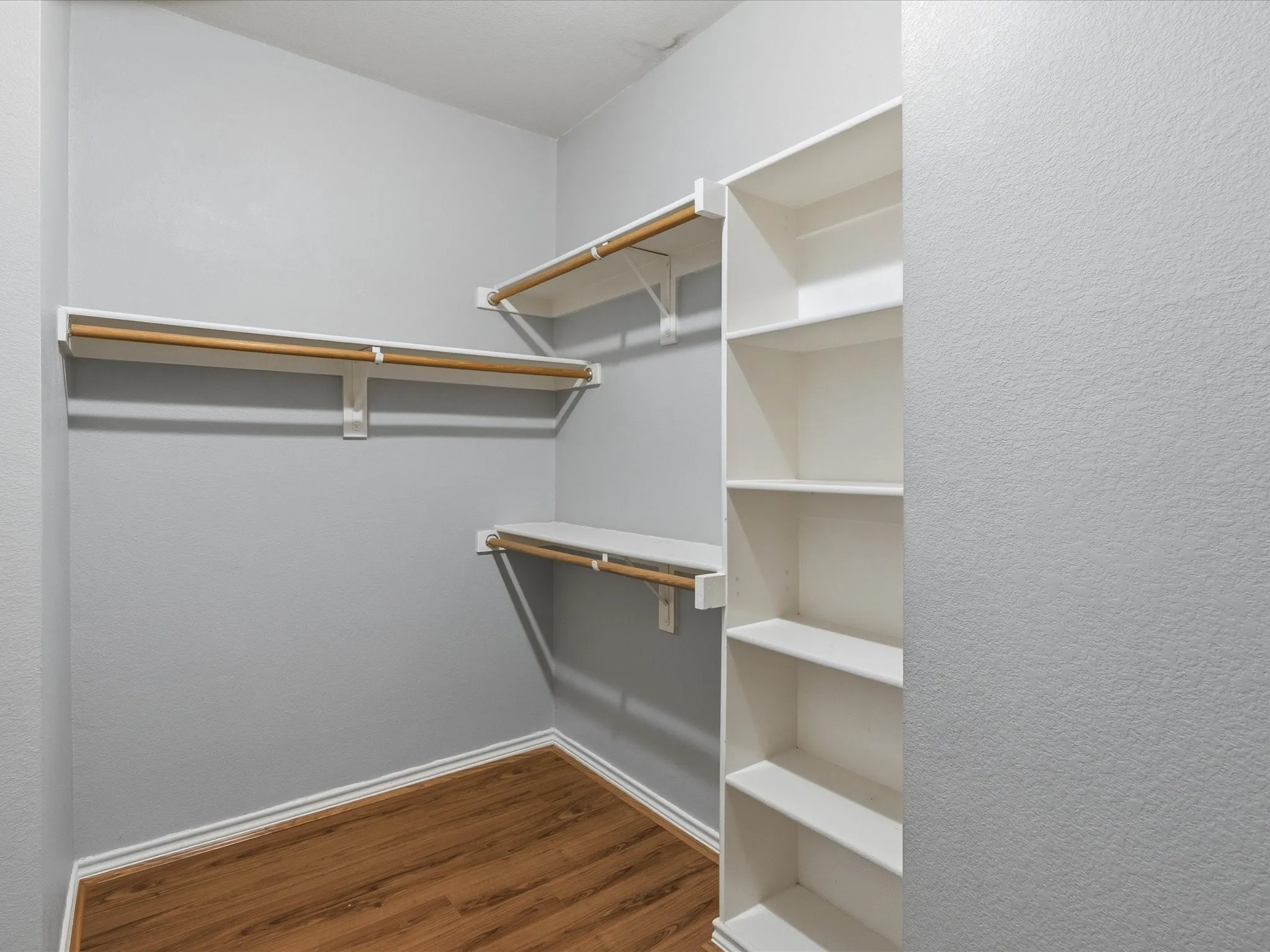 Walk-in closet featuring wood-finish flooring, light gray wall paint, white trim, custom shelving, and clothing rods