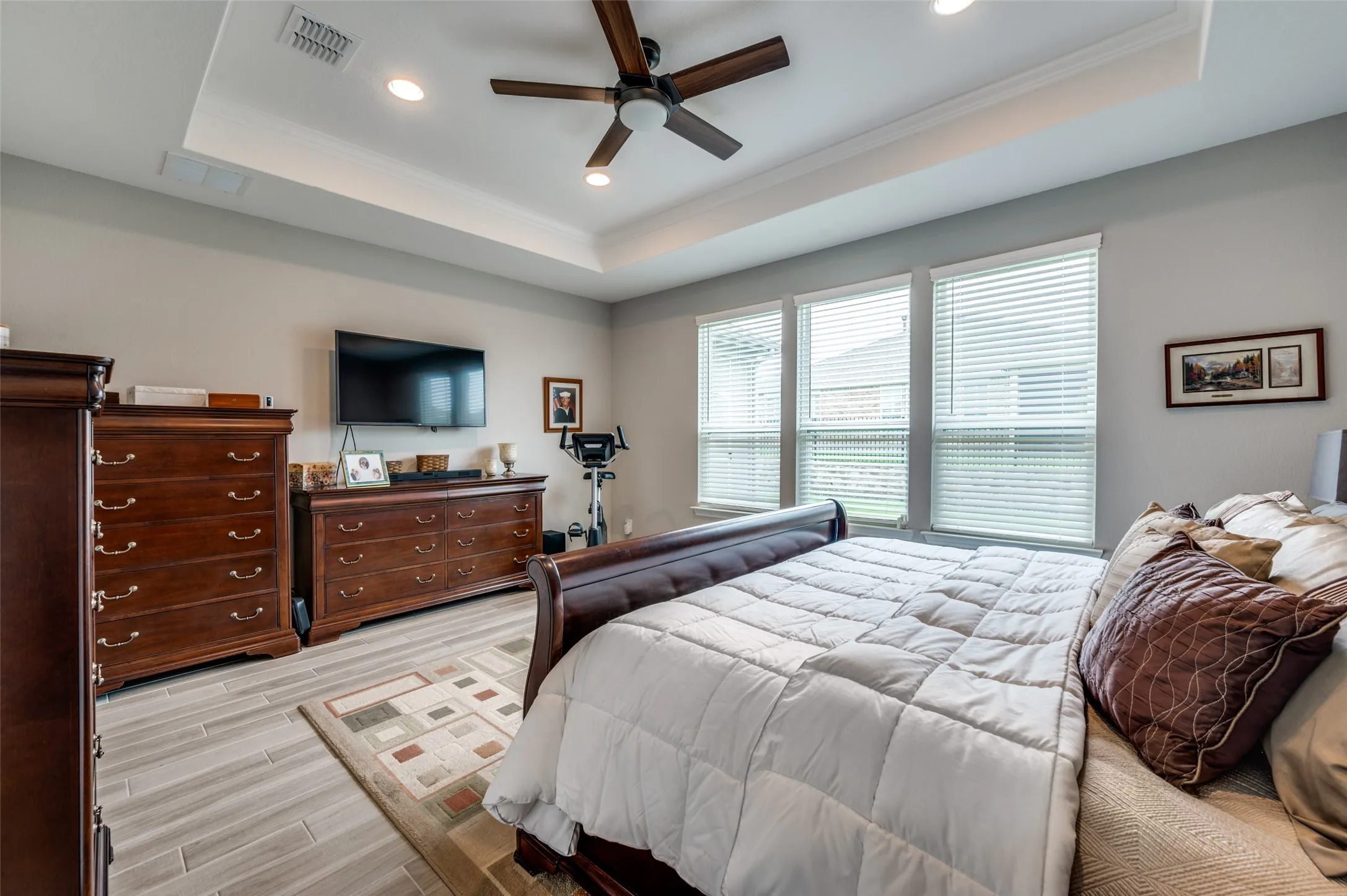 Tray ceiling with recessed lighting and a ceiling fan, light gray walls, triple window configuration with blinds, and wood-finish flooring