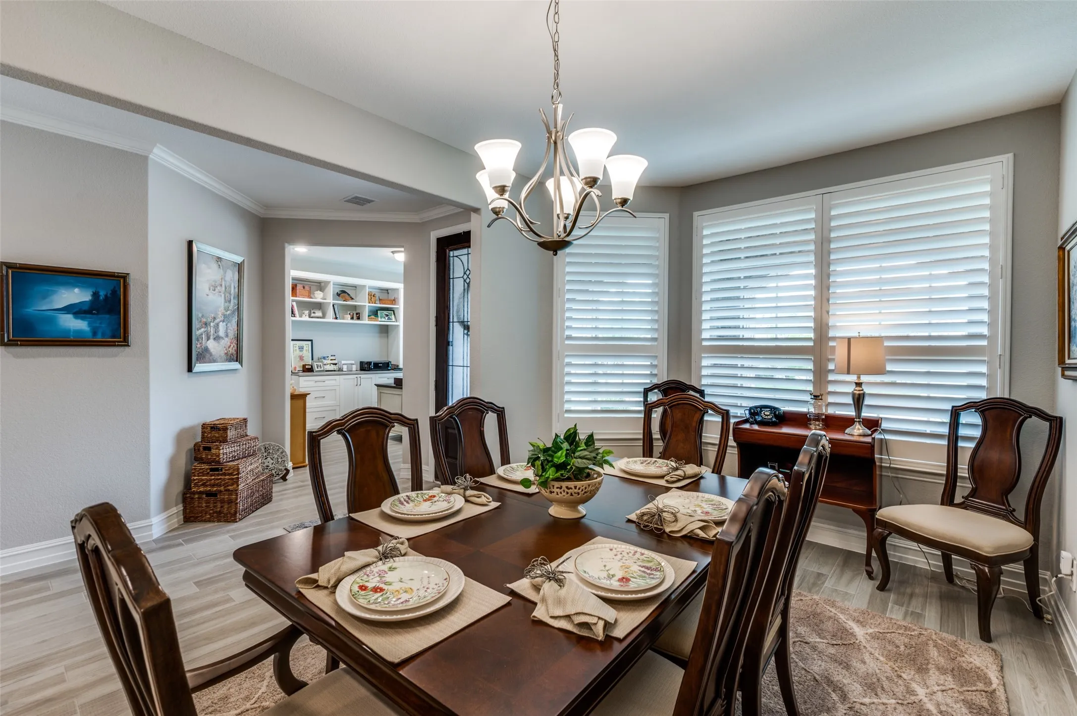 Formal dining area featuring a chandelier, plantation shutters, wood-finish flooring, crown molding, and a visible doorway to an additional space with built-in cabinetry