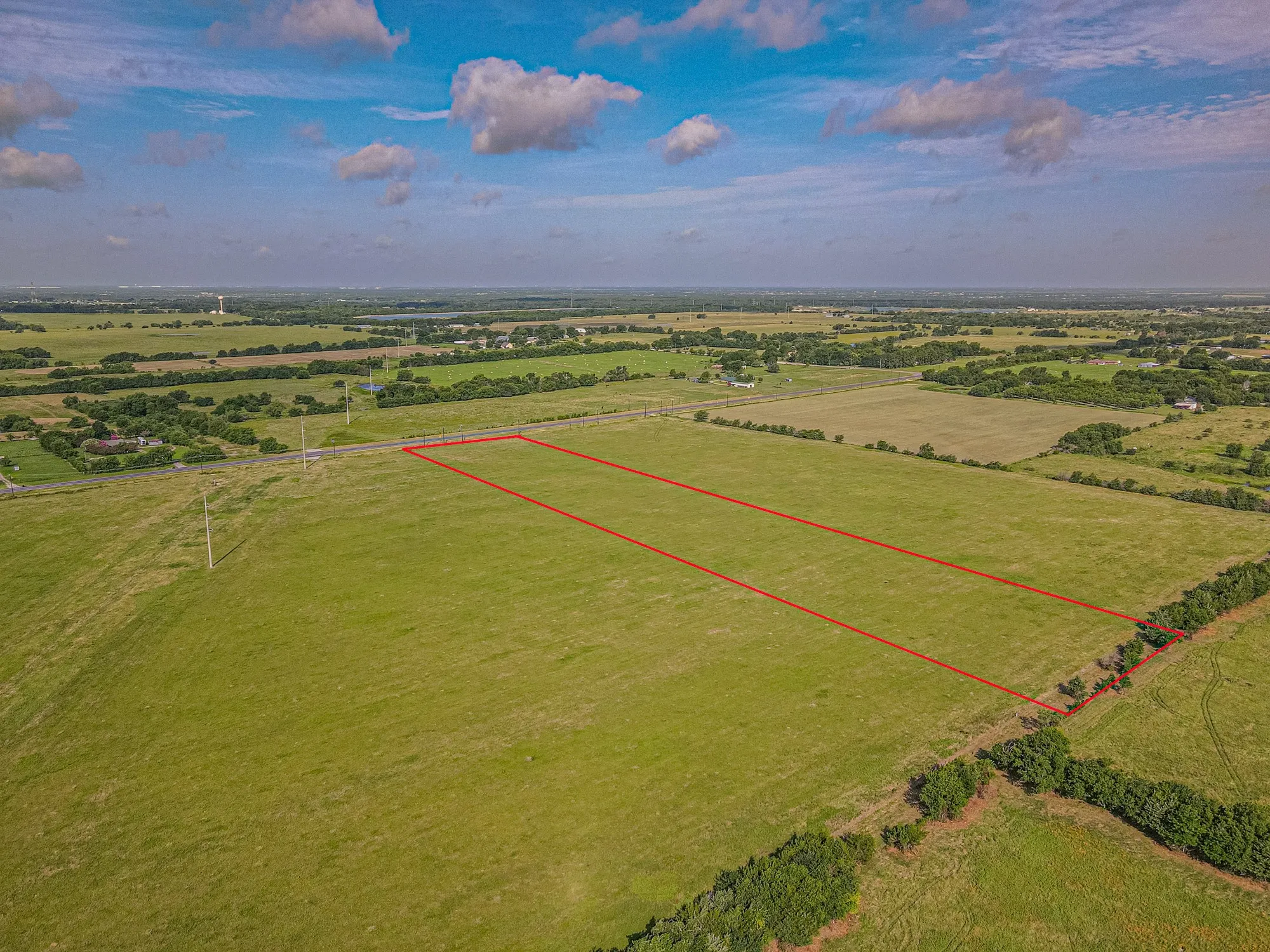 Expansive land parcel featuring a green grass field and clear boundaries, with a distant road and scattered trees