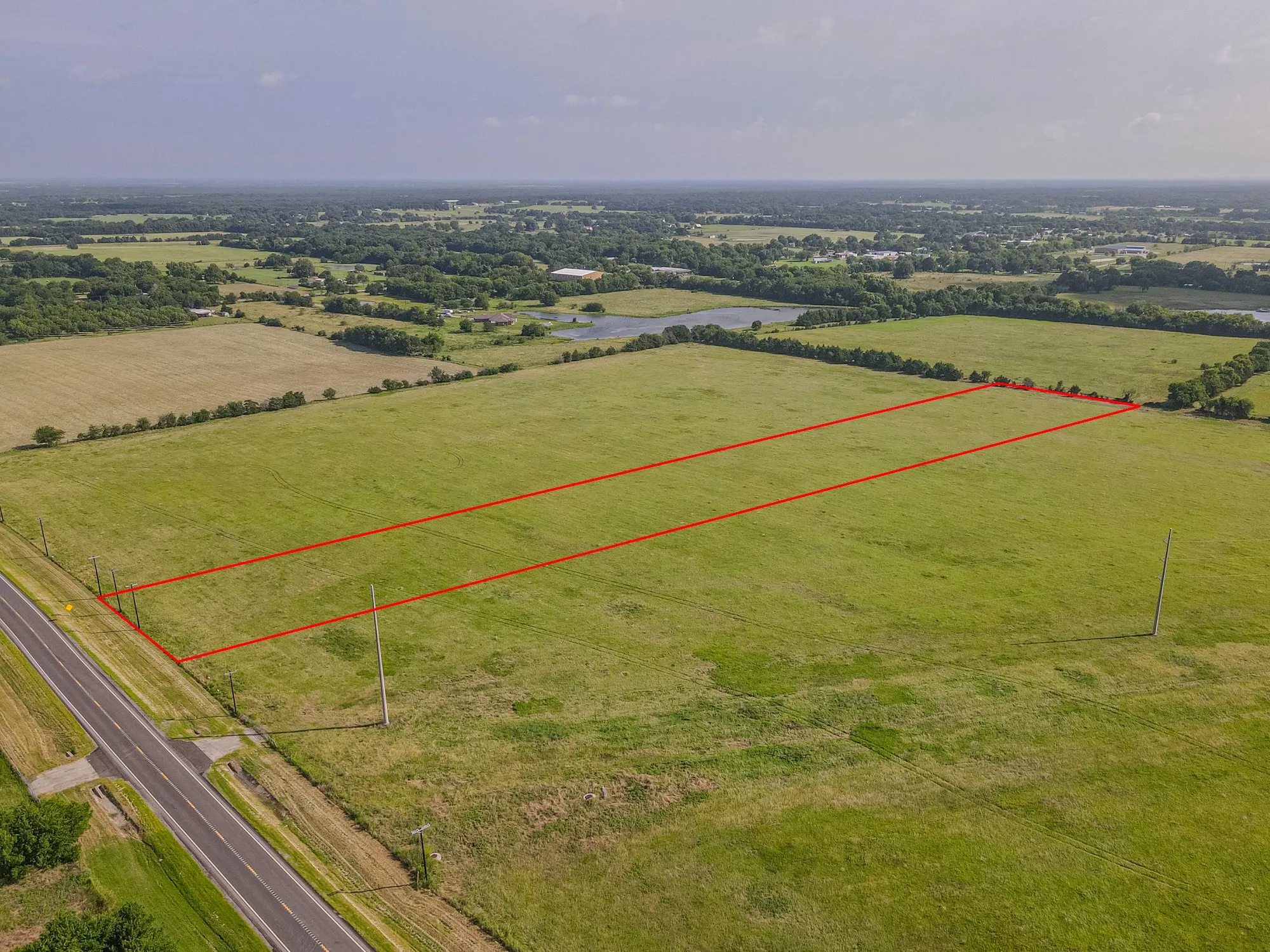 Expansive open land with a clear boundary, featuring a paved road frontage, utility poles, and distant trees