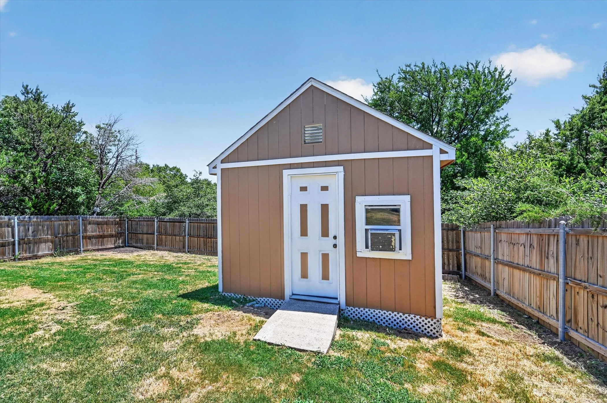 Exterior utility building with a pitched roof, brown siding, a white entry door, a window, and a window-mounted air conditioning unit