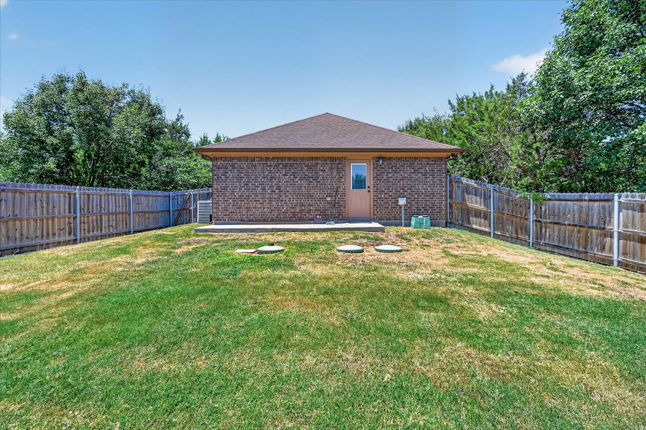 Expansive backyard featuring a privacy fence, a brick exterior with a wood-panel door, a concrete patio, and mature perimeter trees