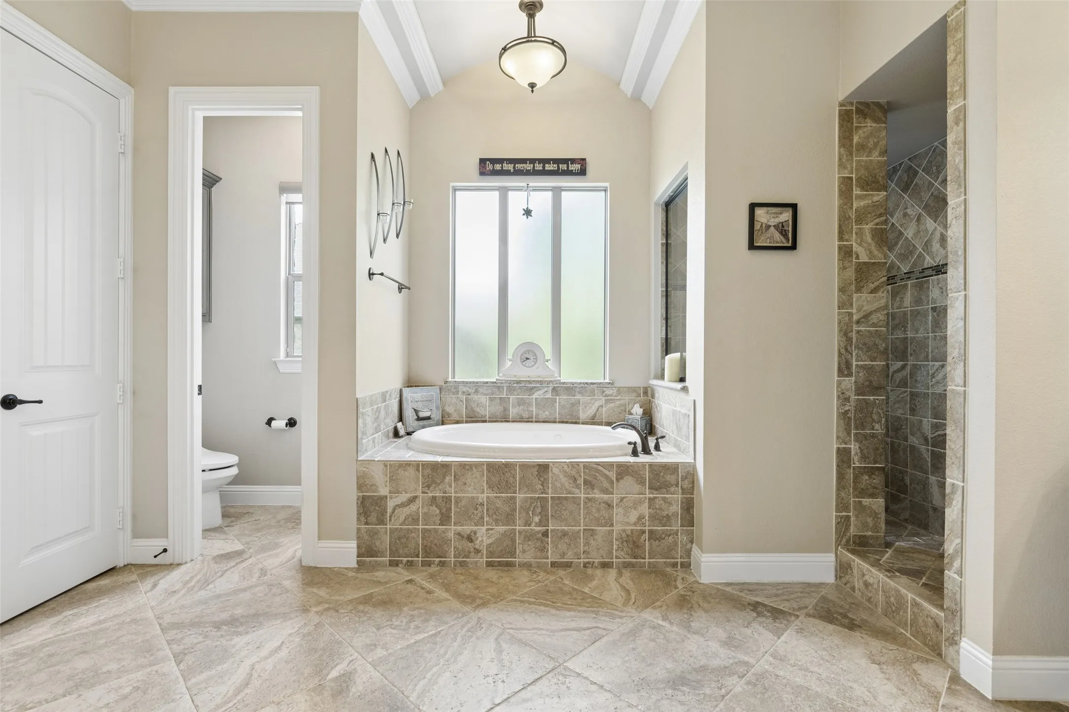 Bathroom featuring a tiled soaking tub, an adjacent walk-in tiled shower with a glass enclosure, vaulted ceilings with crown molding, a decorative pendant light fixture, and large format tile flooring