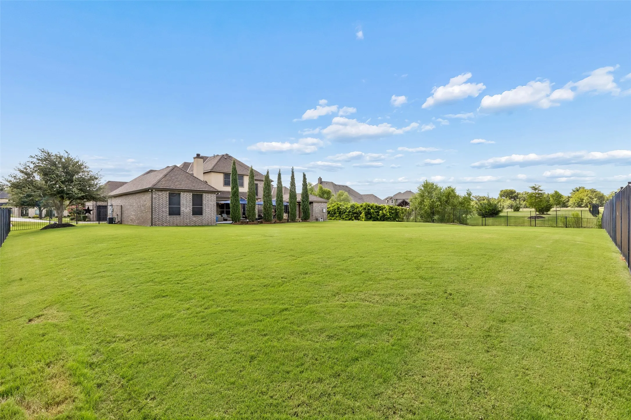 Expansive lawn area featuring a brick exterior home with a multi-gabled roofline, mature trees, and a wooden privacy fence