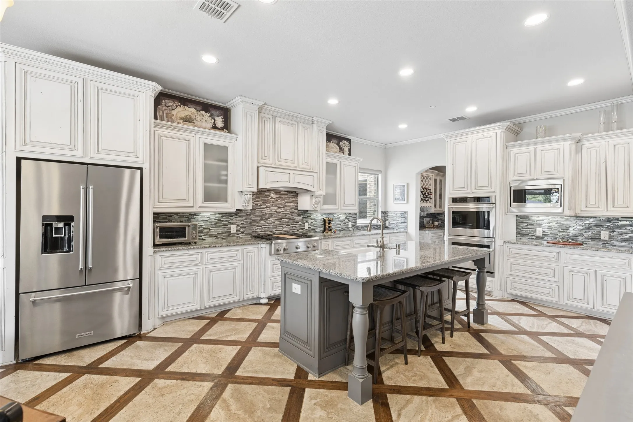 Expansive kitchen featuring a large central island with seating, stainless steel appliances, white cabinetry, granite countertops, and a patterned tile backsplash
